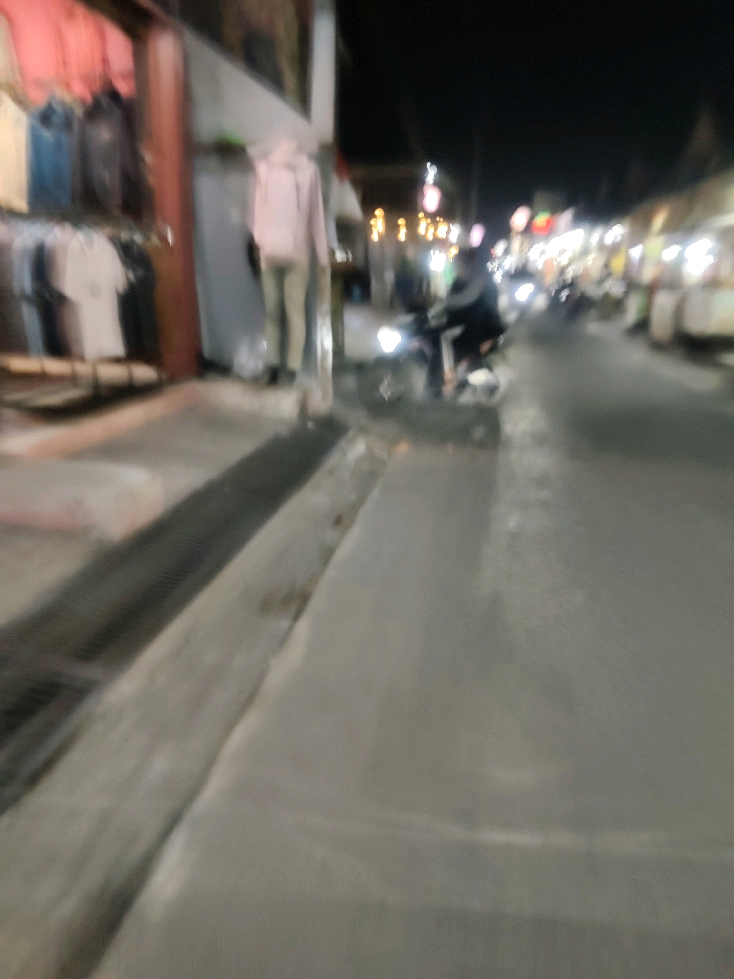 This is a blurred, nighttime photograph taken in Mekarmukti, Indonesia. The image captures a street scene with a storefront on the left and a road with a motorcycle on the right. The storefront displays clothing on racks and a mannequin dressed in a pink hooded garment. The road is wet, suggesting recent rain, and illuminated by streetlights and the headlights of the motorcycle. The overall impression is one of a busy street at night, possibly a market or shopping area.