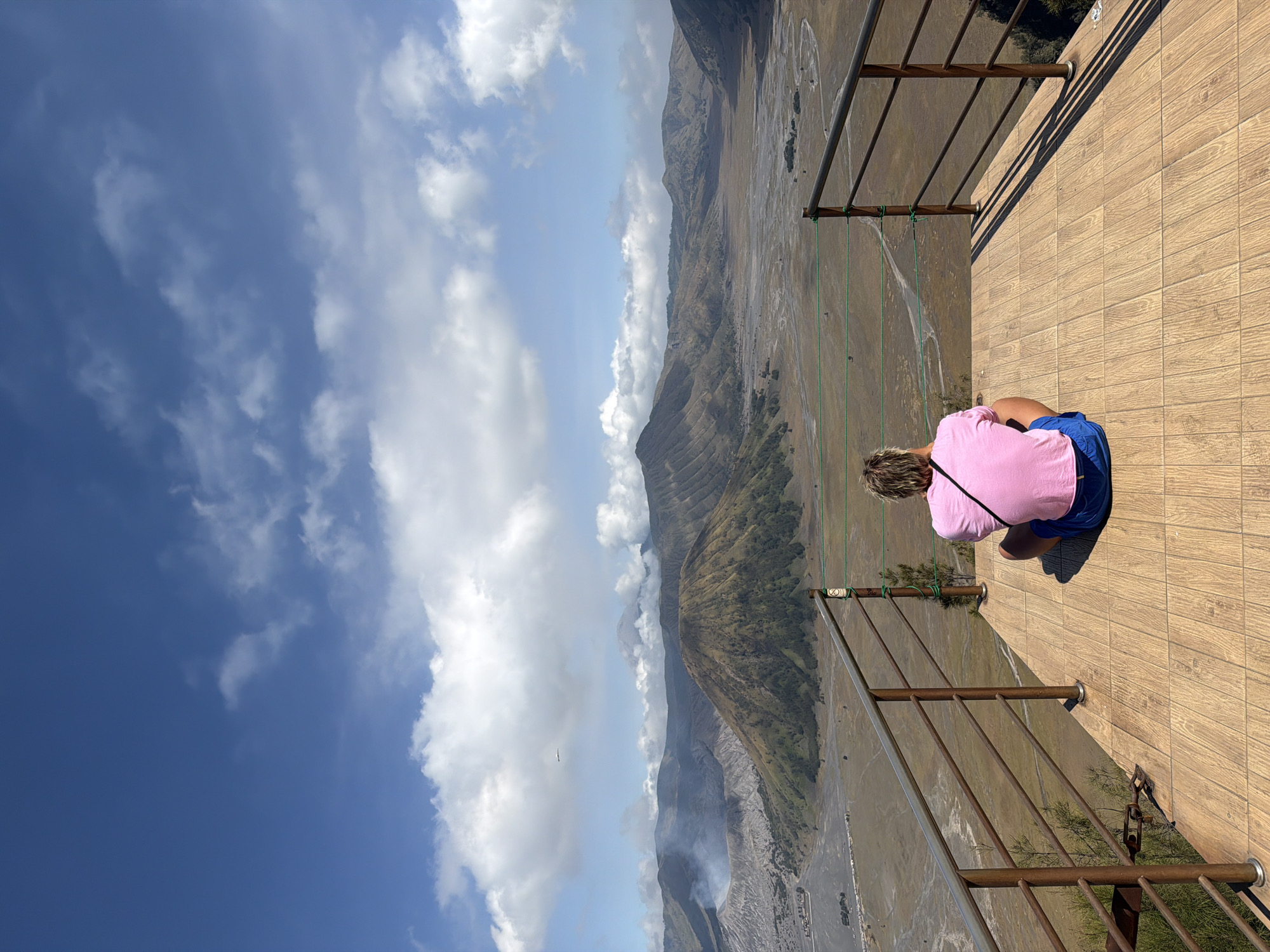 A person wearing a pink shirt and blue shorts is sitting on a wooden platform, facing away from the camera, looking out at a volcanic landscape. The platform has a metal railing. In the background, several volcanoes are visible, with smoke or steam rising from one of them. The sky is blue with scattered clouds. The scene is likely located at a viewpoint in Cemorolawang, Indonesia, overlooking the Bromo Tengger Semeru National Park. The terrain is rugged and mountainous, with a wide, sandy plain in the middle ground.