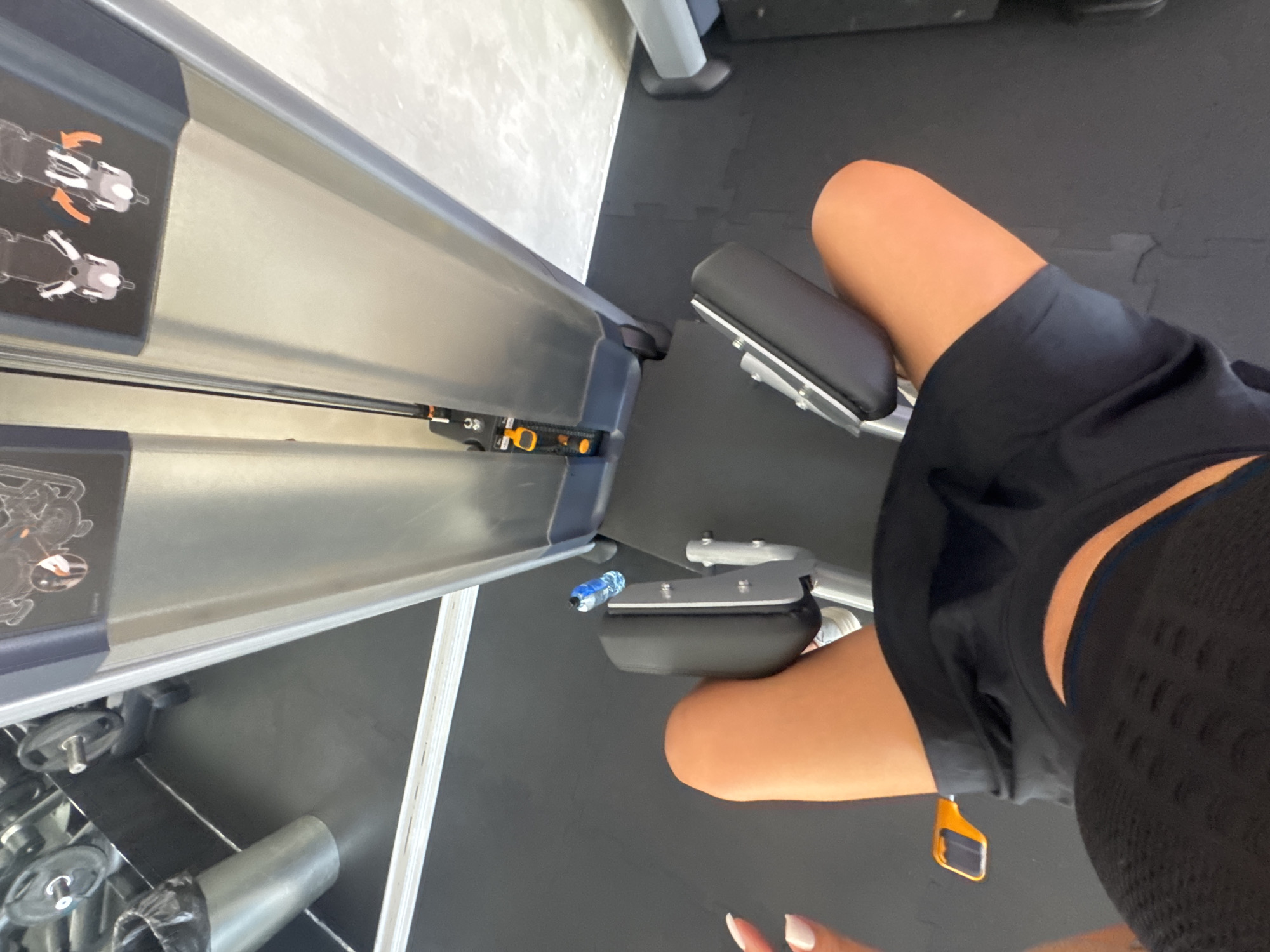 A first-person perspective shot shows a person's legs and torso from a high angle, seated on a weight machine in a gym. The person is wearing black workout shorts and a black top with a mesh detail. They are positioned on the machine, with their knees bent and resting against padded supports. An orange lever is visible on the side of the machine. In the background, a stack of weight plates is partially visible, along with a silver trash can. A water bottle is placed on the floor next to the machine. The floor is covered with black mats. The gym appears to be located in Budva, Montenegro, as indicated by the metadata.