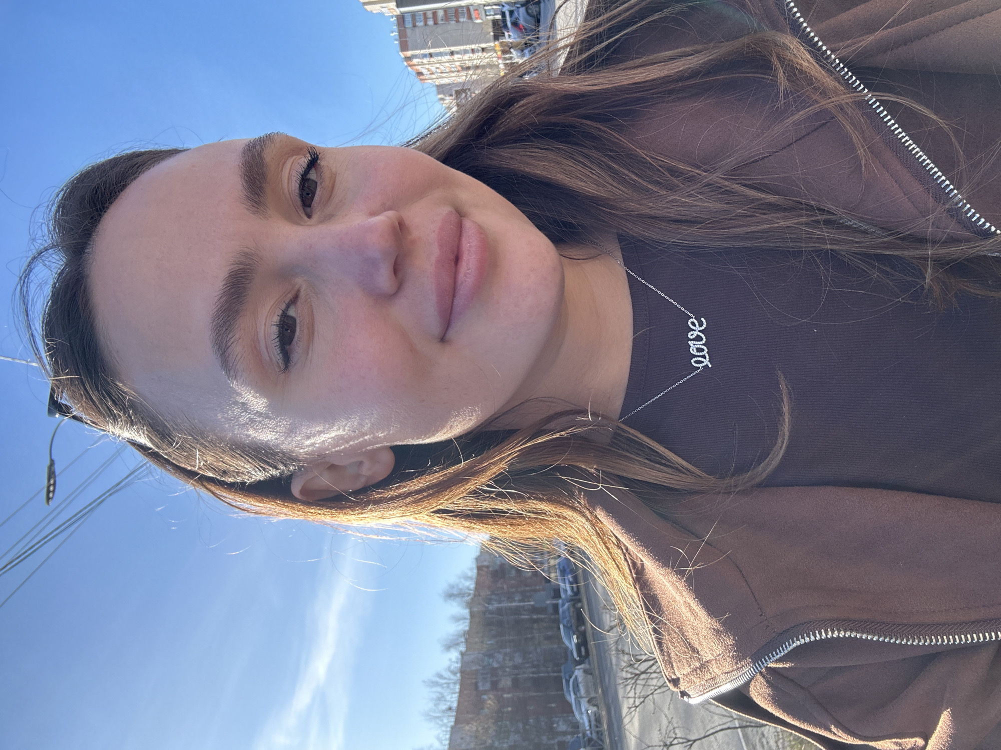 A woman is taking a selfie outdoors on a sunny day. She has fair skin, brown hair, and is wearing a brown top and jacket. She has a silver necklace with the word "love" on it. Behind her, there's a clear blue sky and some buildings, possibly in an urban area. The precise location in Surgut, Russia is not discernable from the image.