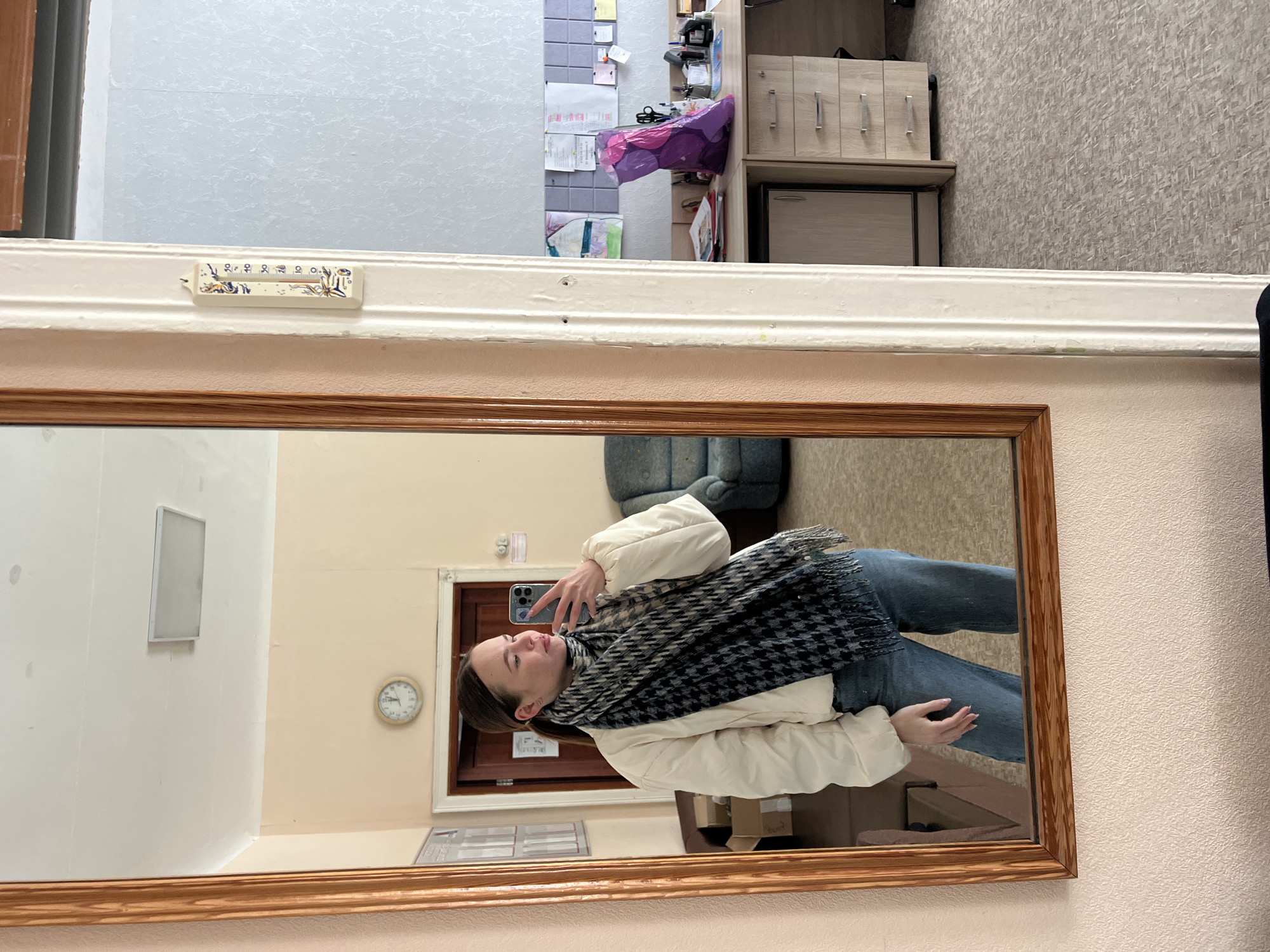 A person is reflected in a mirror, presumably taking a selfie. The person is wearing a white jacket, blue jeans, and a dark scarf. The background in the mirror shows a room with a door, a clock, and an office chair. Outside the mirror's reflection, one can see a portion of a wall, a thermometer attached to the wall above the mirror, and an out-of-focus cabinet and other items in the upper portion of the frame. Based on the information available, it's impossible to determine the exact location in Chaikovsky, Russia.