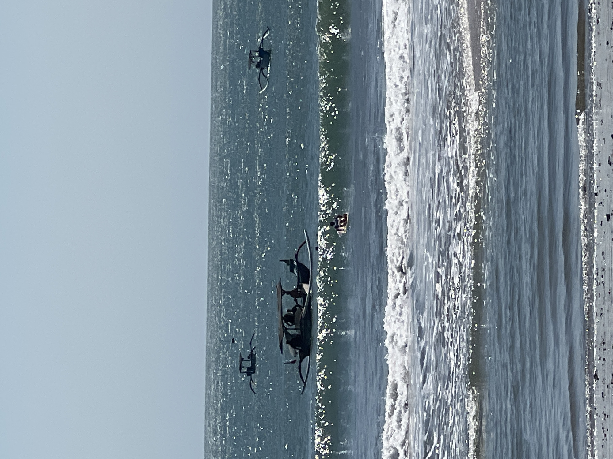 The image depicts the ocean off the coast of Kuta, Indonesia, with several traditional outrigger boats visible on the water. The sky is a pale blue and hazy, suggesting a bright, sunny day. The sea is a deep blue, with whitecaps forming on the waves. In the foreground, the waves are breaking closer to the shore, with some white foam visible. In the mid-ground, a larger outrigger boat with a canopy is carrying several people. Further out to sea, two smaller outrigger boats are also visible. One person is on a surfboard or boogie board in the water between the larger boat and the breaking waves. The shore in the very bottom of the image appears to be sandy with some small rocks or pebbles.
