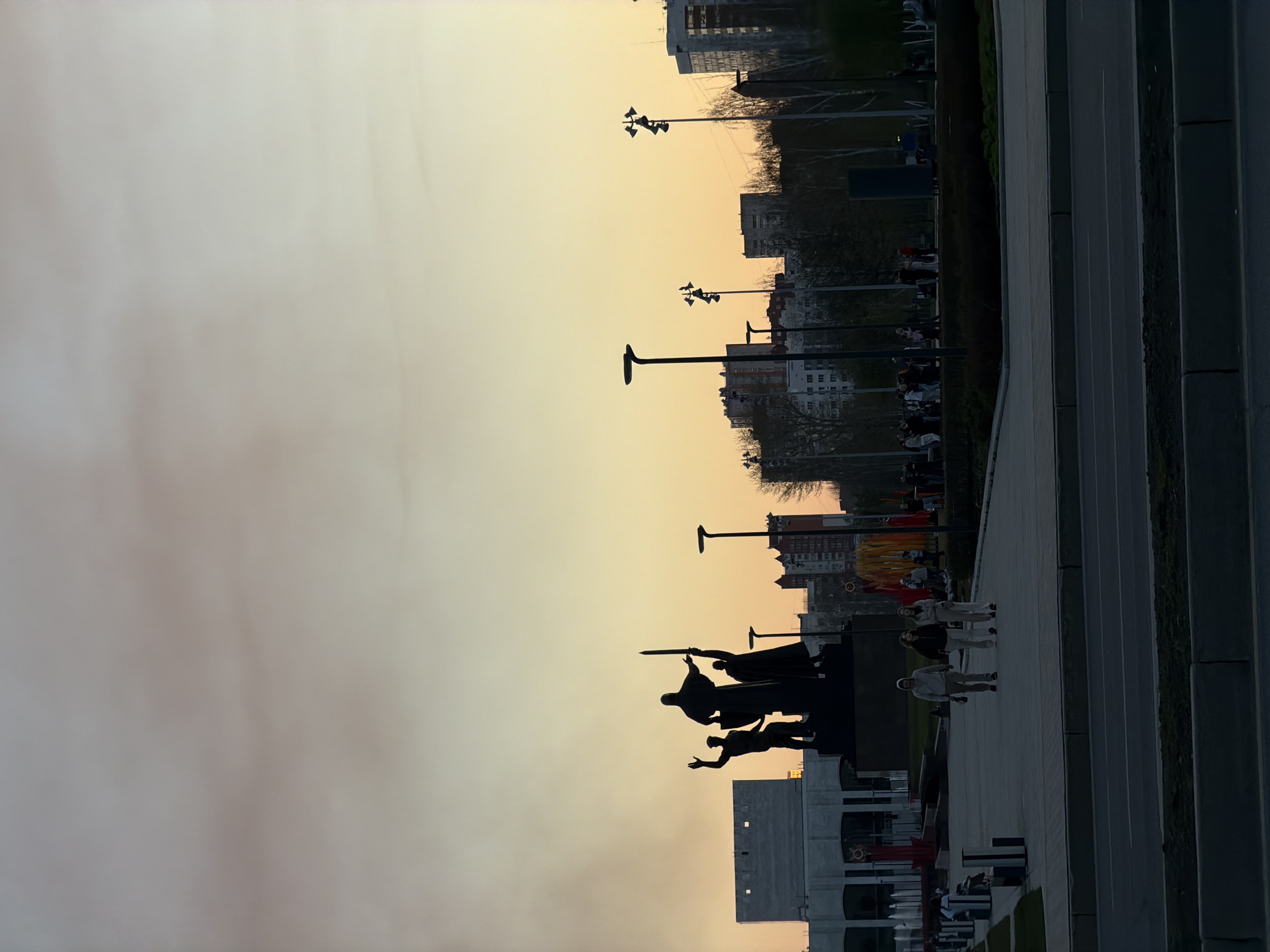 The image depicts a cityscape at sunset, likely in Perm, Russia, based on the presence of the "Front to the Rear" monument.
The "Front to the Rear" monument is silhouetted against the colorful sky. Several streetlights are visible along the path. A group of people are walking on the paved area next to the monument. Tall buildings are present in the background.