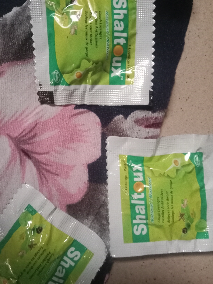 Within the city of Iyiowa Odekpa, Nigeria, an overhead view captures several individually packaged "Shaltoux" brand cough lozenges resting on a patterned fabric surface. Three white, rectangular foil sachets are visible, each featuring a green central panel with the brand name "Shaltoux," text indicating "Cough Lozenges," "Natural / Naturel," "Pastilles Antitussives," "Relieves sore throat," and "Soulage les maux de gorge," along with a depiction of a round orange lozenge and herbal illustrations. One sachet specifies "1 Lozenge / 1 Pastille." The sachets exhibit serrated edges along their seals. The underlying surface is a dark-colored fabric, possibly black or dark brown, patterned with large, stylized pink floral elements, specifically prominent petals. No individuals or specific actions are depicted.