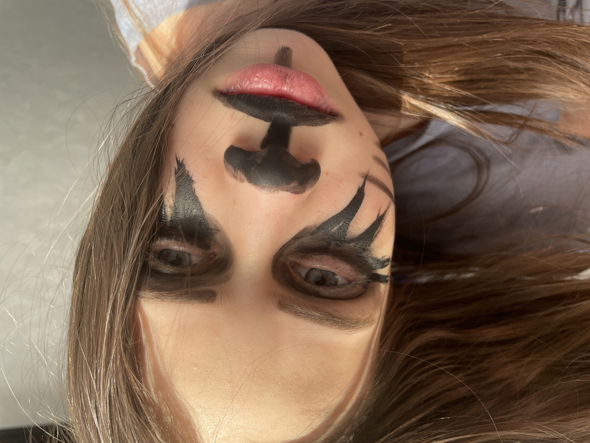 A close-up portrait of a young person with brown hair, turned upside down. Their face is decorated with black paint, resembling a stylized animal or theatrical mask. The paint covers their lips, nose, and around their eyes, creating sharp, wing-like shapes. The background is indistinct. There is no information to indicate this image was taken in Izhevsk, Russia.