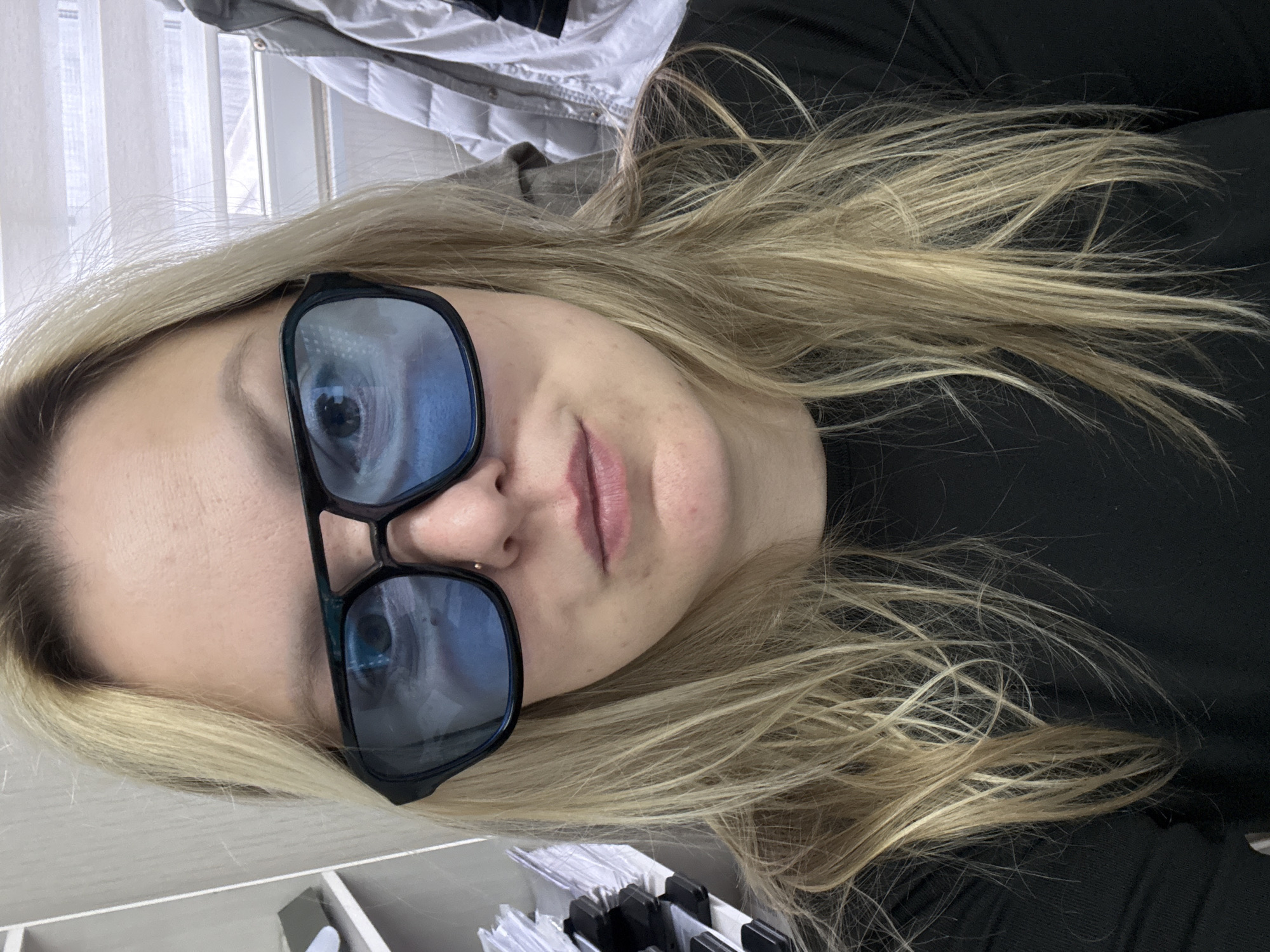 A woman with blonde hair is wearing dark-framed glasses. She is wearing a black shirt. There is a white jacket visible in the background. The objects in the foreground appear to be stationary. The location in the city of Noviy, Russia is not discernible from the image.