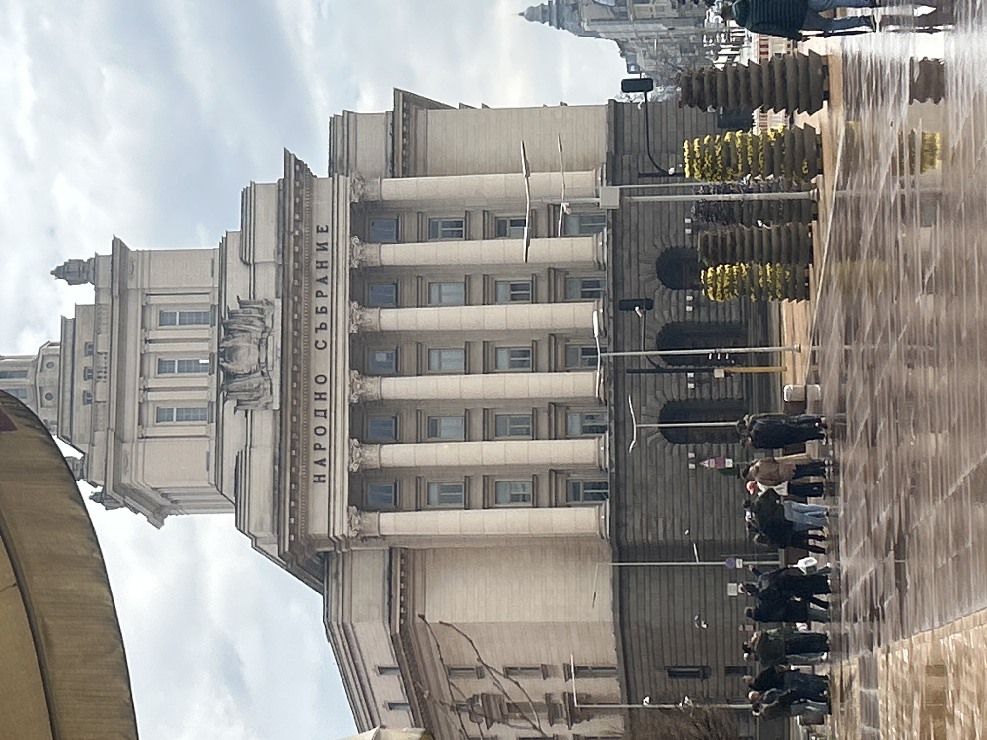 The image depicts the National Assembly building in Sofia, Bulgaria. The building is large, with a white facade, columns, and several windows. A sign on the side of the building reads "НАРОДНО СЪБРАНИЕ," which translates to "National Assembly." In the foreground, there is a fountain or wet pavement reflecting the building and surrounding objects. Several pedestrians are walking by the building. There are also some planters with plants in front of the building. The sky is overcast.