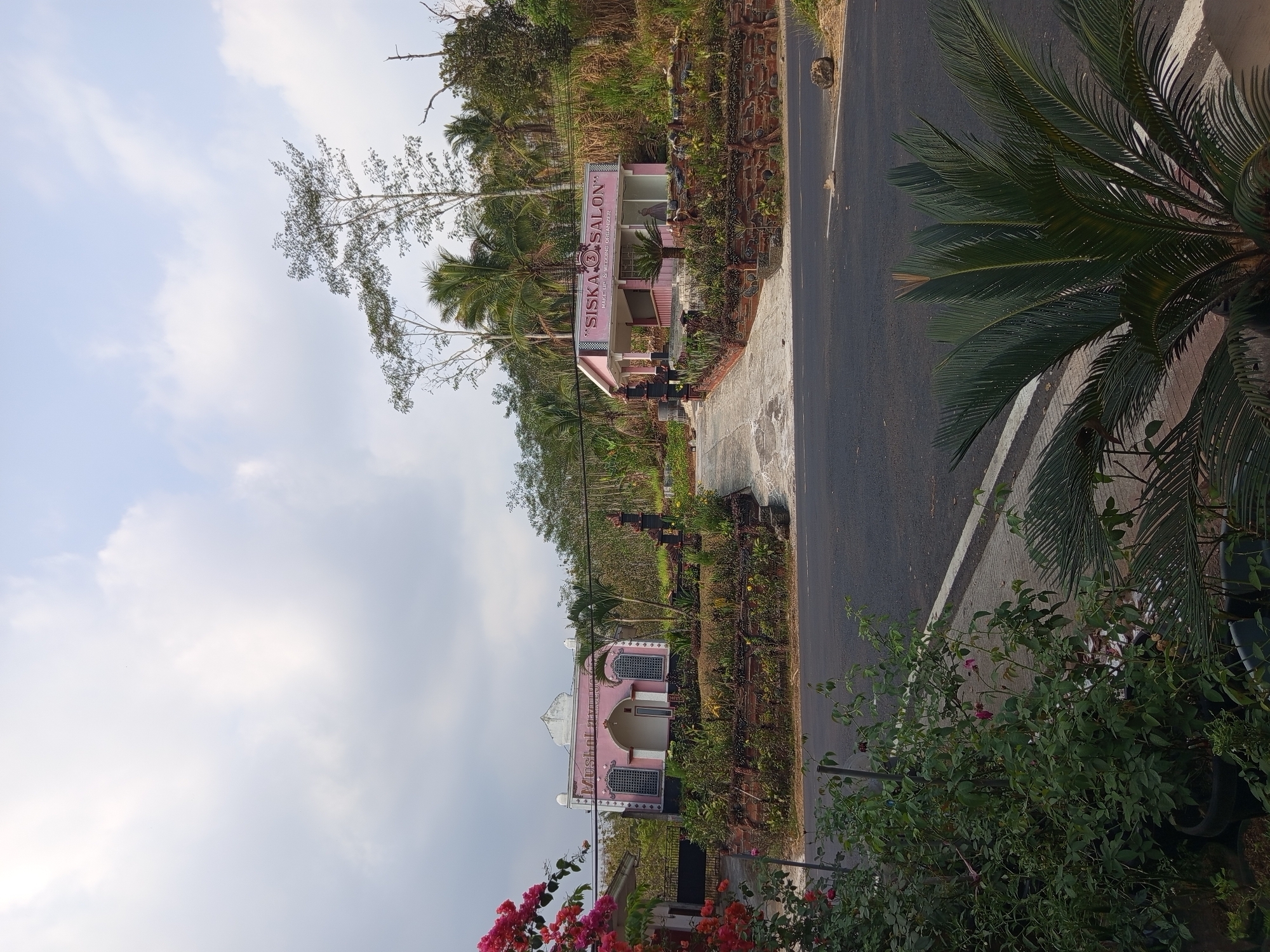 The image captures a roadside view in Sumberpucung, Indonesia, featuring two distinct pink structures. On the left, a building displays text that appears to read "MusHoL LauWud." This structure has arched windows with decorative grilles and a white-domed roof. To the right, a smaller, open-air pavilion is labeled "SISKA SALON MAKE UP & WEDDING ORGANIZER." Both buildings are situated amidst lush greenery, with flowering plants and trees forming a natural backdrop. A paved road runs in front of the buildings, and a portion of a palm-like plant is visible in the foreground. The sky is overcast with scattered clouds.