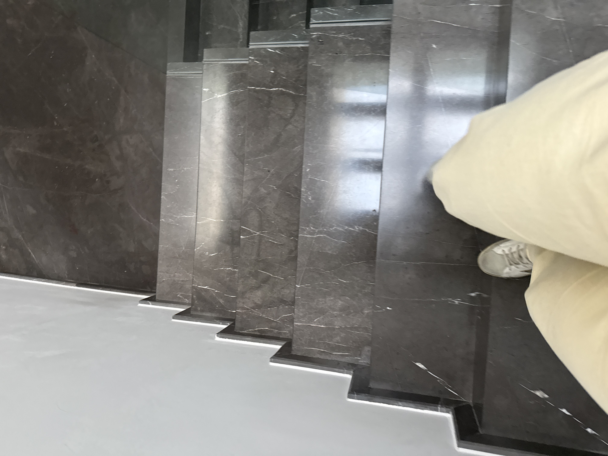 A person in beige trousers and white sneakers walks down a set of dark gray marble stairs in San Bernardino Tlaxcalancingo, Mexico. The staircase is flanked by a gray marble wall and a white floor. The bottom edges of the stair treads are illuminated.