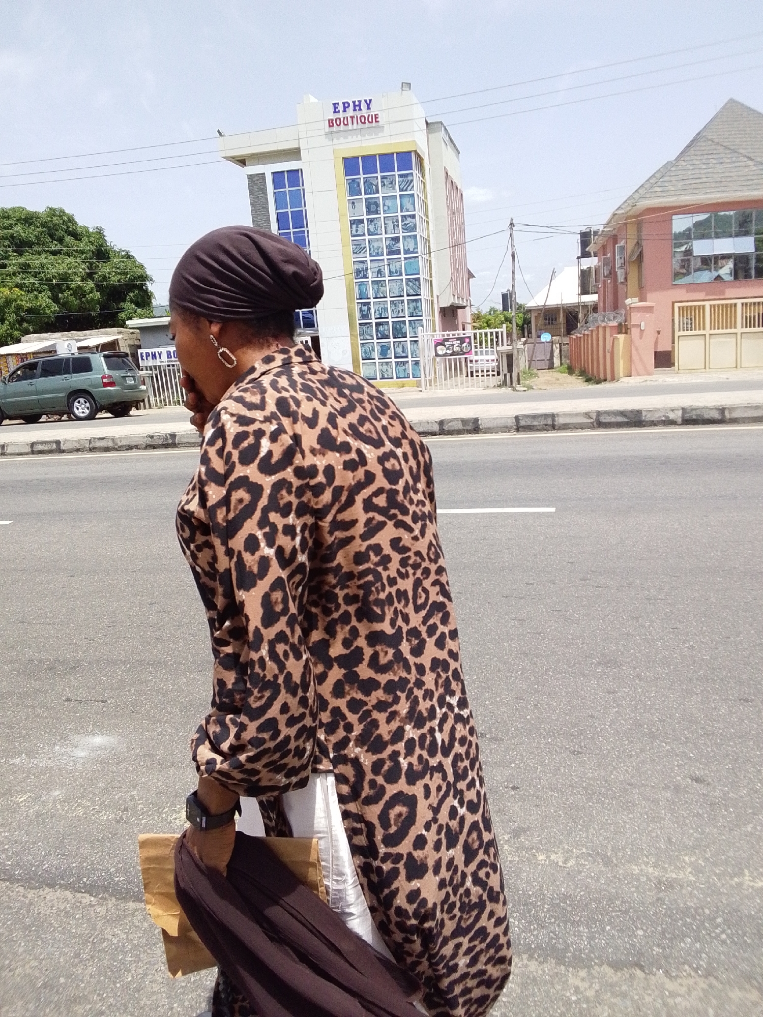 The image presents a daytime scene of a person on an urban street in Adankolo, Nigeria. The subject, viewed from a side-rear angle, is wearing a prominent brown and black leopard print long-sleeved top and a dark brown head covering. They have large, oval-shaped earrings and a dark watch or band on their right wrist. In their right hand, they carry a brown paper bag and a piece of dark fabric. The person appears to be walking or standing beside a paved road.
In the background, a modern, multi-story building with a white and gold facade and distinctive blue-tinted windows stands prominently. The name "EPHY BOUTIQUE" is clearly visible in red and blue lettering on its top section. To the right of this building, another two-story structure painted peach or pink, featuring a grey tiled roof and reflective windows, is visible. On the far left, behind a white fence, a green SUV (likely a Toyota Highlander) is parked.
The scene includes an asphalt road with a visible white marking line, a concrete curb, and a sidewalk area. Overhead utility poles and wires are visible against the bright, clear sky, indicating sunny weather. A smaller sign near the white fence also bears the partial text "EPHY BO...". The overall setting suggests a commercial or mixed-use area.