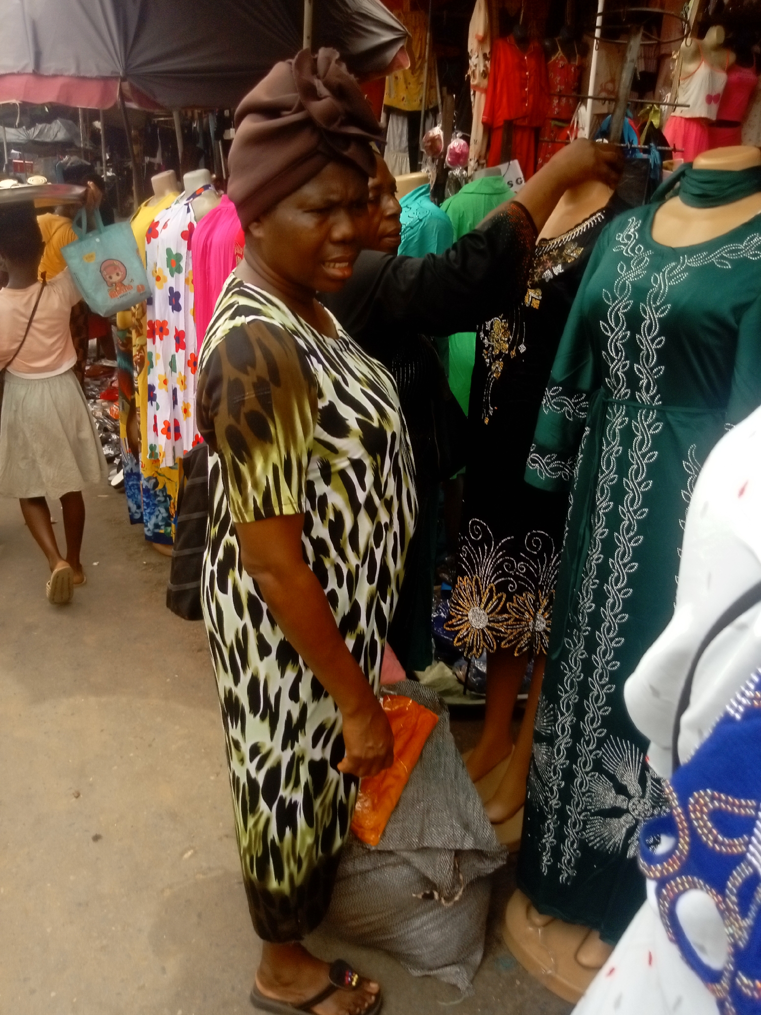 A medium shot taken at ground level shows a woman with a brown head wrap standing in a marketplace or outdoor vendor area in Fegge, Nigeria. She is wearing a patterned white, black and olive green short-sleeved dress. In front of her is a large gray burlap sack with a folded orange cloth on top. She is holding the cloth in her right hand. She is wearing dark sandals. Behind her are mannequins dressed in colorful dresses. A vendor's hand and arm is reaching out to adjust the dress on one of the mannequins. In the background, other people are walking through the area. A light blue tote bag with a cartoon on it is being carried by one of the people in the background. The scene appears to be taken during the daytime, under an awning.