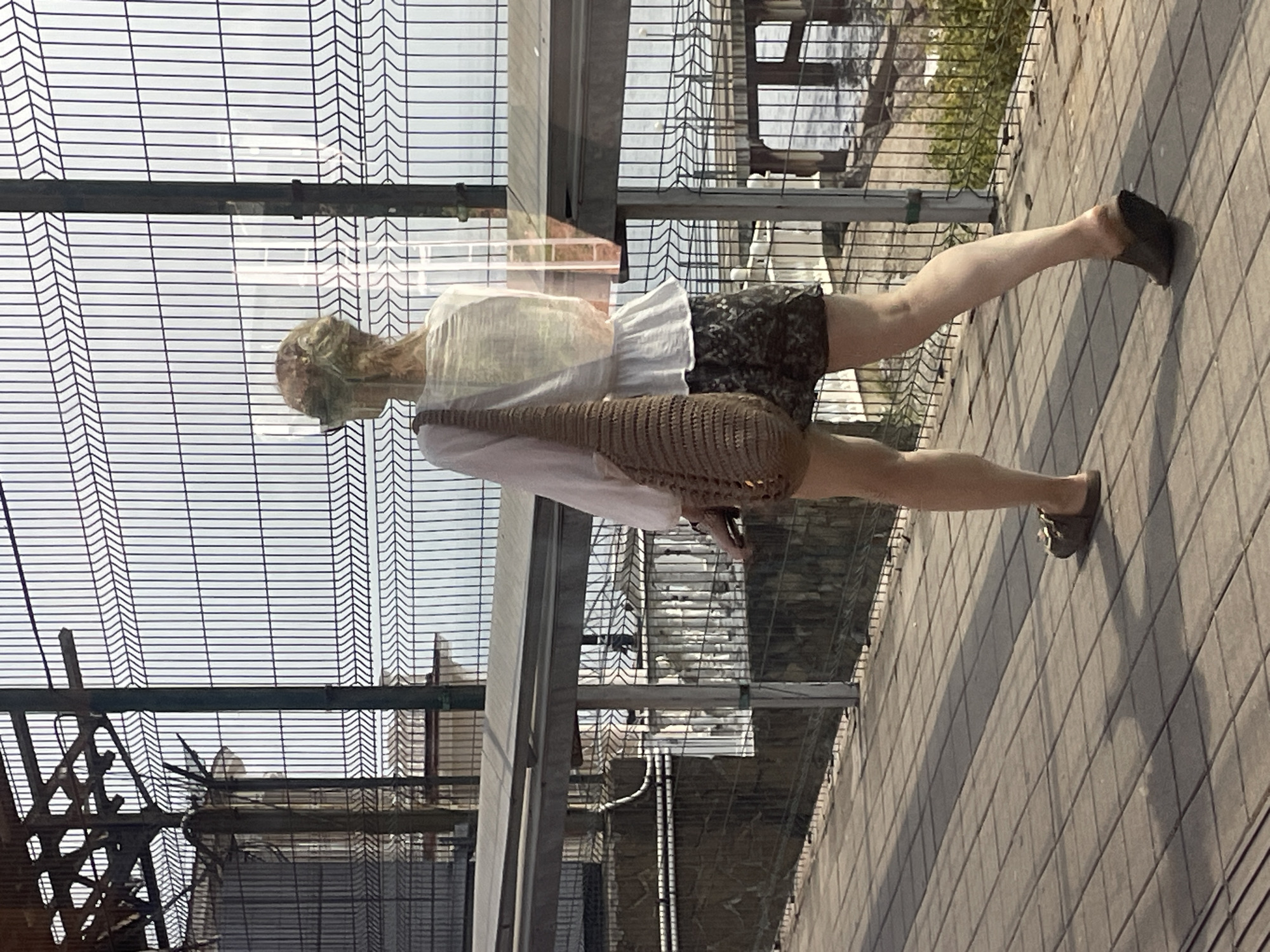 The image shows a woman walking on a paved walkway near a wire fence with the sea in the background. The woman is wearing a white blouse, patterned shorts, and brown sandals. She is carrying a brown crocheted bag over her left shoulder. The walkway is made of large, square concrete tiles, and shadows are cast across it from the fence and an overhead structure. In the background, there is a metal pier extending into the water. The wire fence has vertical and horizontal elements, with some metal poles supporting it. The overall scene appears to be an outdoor public area, possibly a promenade or boardwalk, consistent with locations found in the city of Sochi, Russia, which is known for its coastal resorts.