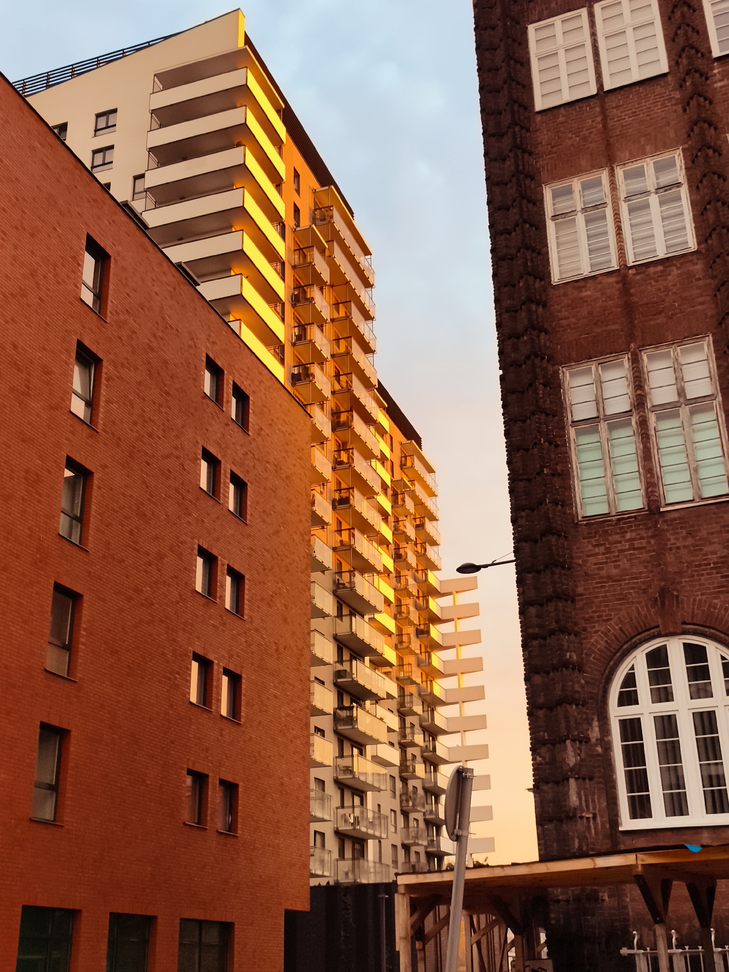 This image shows a view of modern and older buildings against a bright sky, likely at sunset. On the left is a building with a brick facade and several windows. To its right is a tall, slender apartment building with many balconies, bathed in warm, golden light. On the far right is a darker, older building made of red brick, featuring large, white-paned windows, including a prominent arched window at the bottom. A streetlamp is visible extending from the older building towards the apartment building. The scene is located in the city of Gdańsk, Poland, specifically in the area of the former shipyard, which is now a rapidly developing residential and business district. No people are visible in the frame.