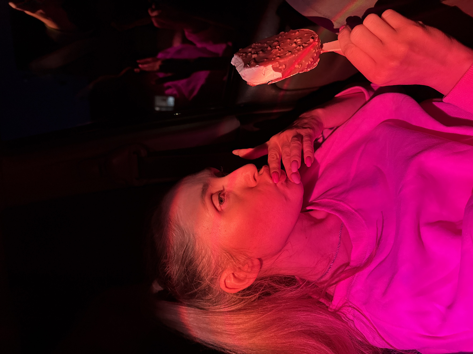 A person is lying down, holding an ice cream bar. The person is wearing a pink sweater. The ice cream bar has a chocolate coating with nuts. The person's hair is blonde. The setting is dark with a pink light illuminating the person and the ice cream. The person is in Chaikovsky, Russia.
