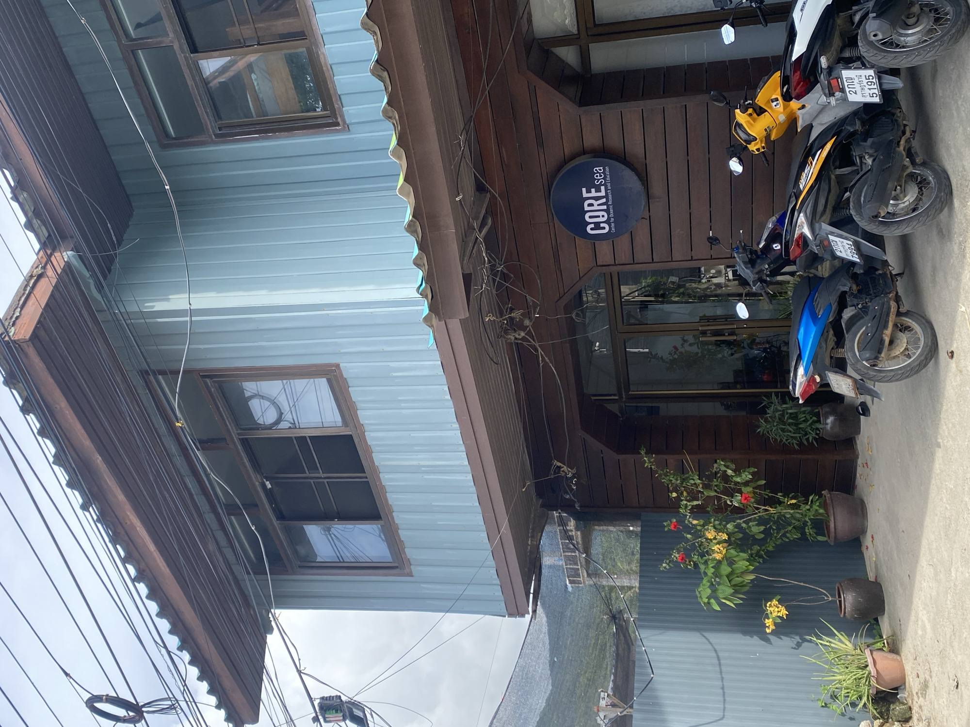 Here's a description of the image:
The photo captures a street view in Baan Chaloklum, Thailand, showcasing a two-story building. The first story is clad in wood and has a sign that reads "CORE sea Center for Oceanic Research and Education". There is a glass door storefront. A light blue second story has visible windows. The building has a brown corrugated metal roof.
Parked directly in front of the entrance are two motorbikes, one blue and one yellow and white. Flower pots with plants are lined up near the entrance. Power lines are visible crossing above the building.