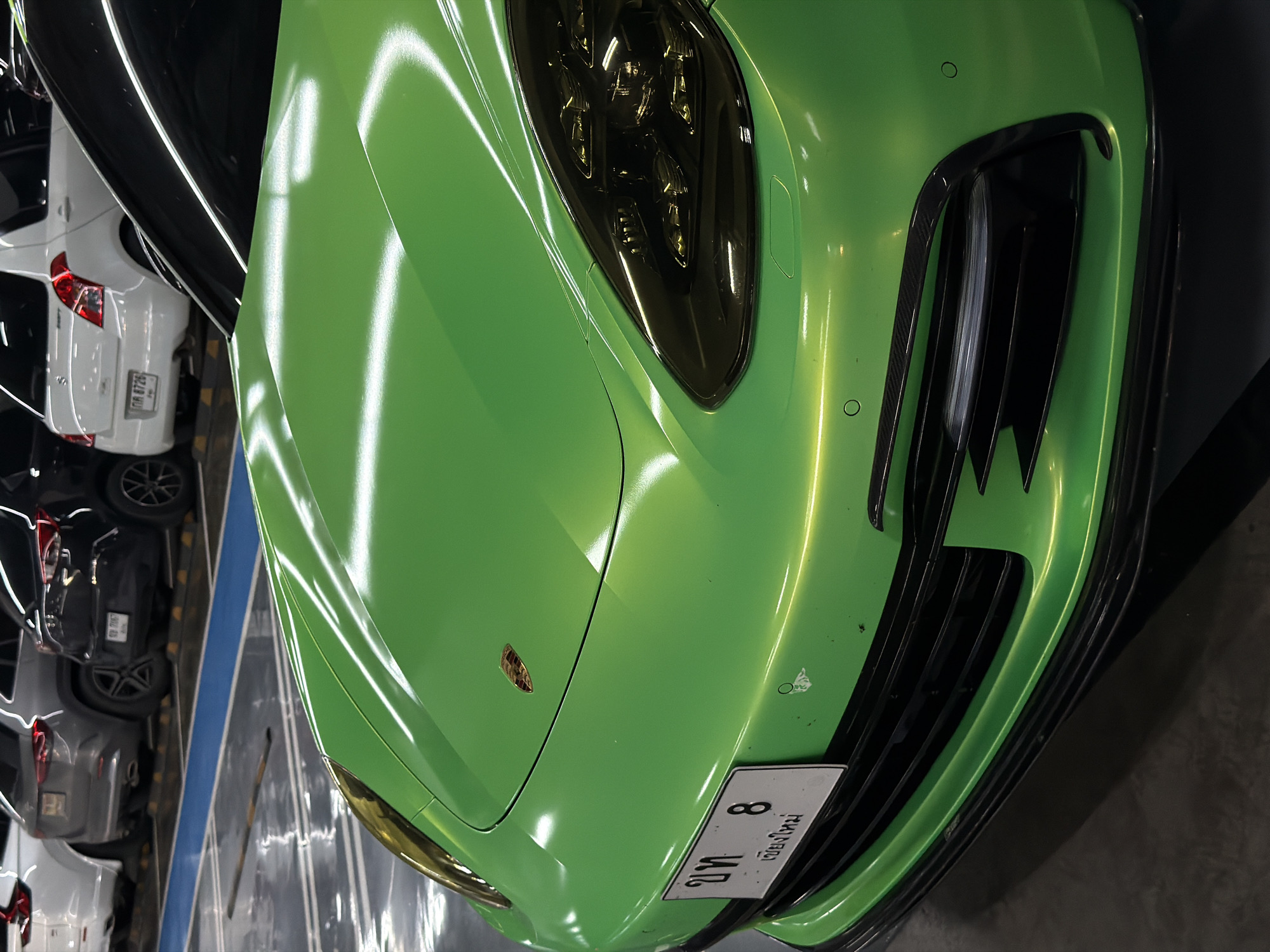 A bright green Porsche Panamera is parked in an indoor parking lot, likely in the city of Nong Pa Khrang, Thailand. The car's hood is prominently displayed, reflecting the overhead lighting. The front bumper, grille, and a portion of the headlight are also visible. A white license plate with Thai characters and the number "8" is affixed to the front of the car. In the background, other vehicles are parked in designated spots, including a white hatchback, a grey SUV, and a black sedan. The parking lot floor appears to be a polished concrete or similar surface, marked with blue and yellow lines. The lighting suggests it is daytime, but the artificial light from the parking lot is dominant. There are no people visible in the frame.