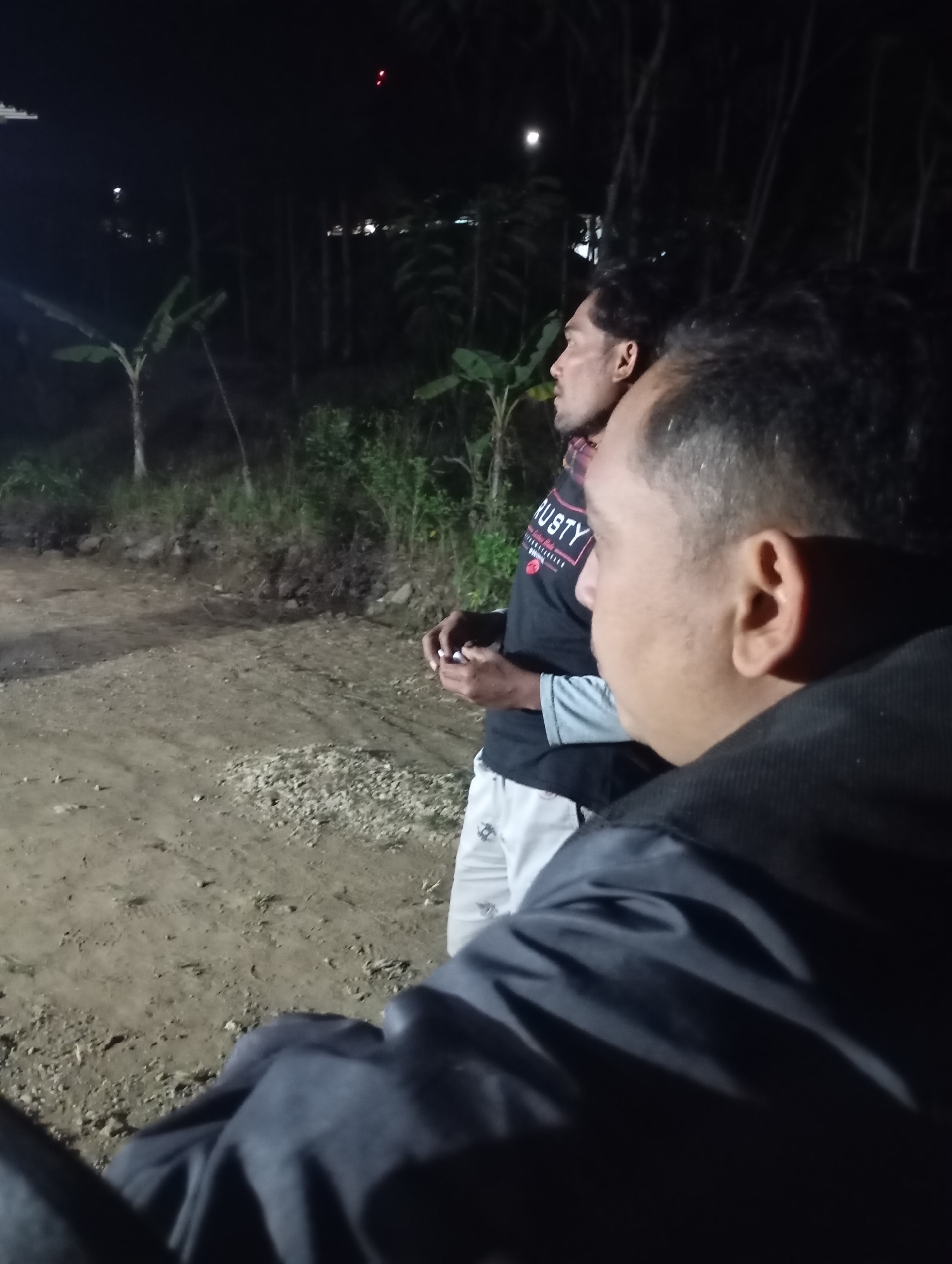 In Ketepung, Indonesia, at night, two men stand on a dirt road. The man on the left faces away from the camera, looking to the right. He wears a black t-shirt with red and white text that reads "RUSTY" and light-colored pants. The man on the right faces the same direction as the other man, with his back to the camera. He wears a dark jacket. The environment is dimly lit, with trees and foliage visible in the background. A red light is visible in the distance.