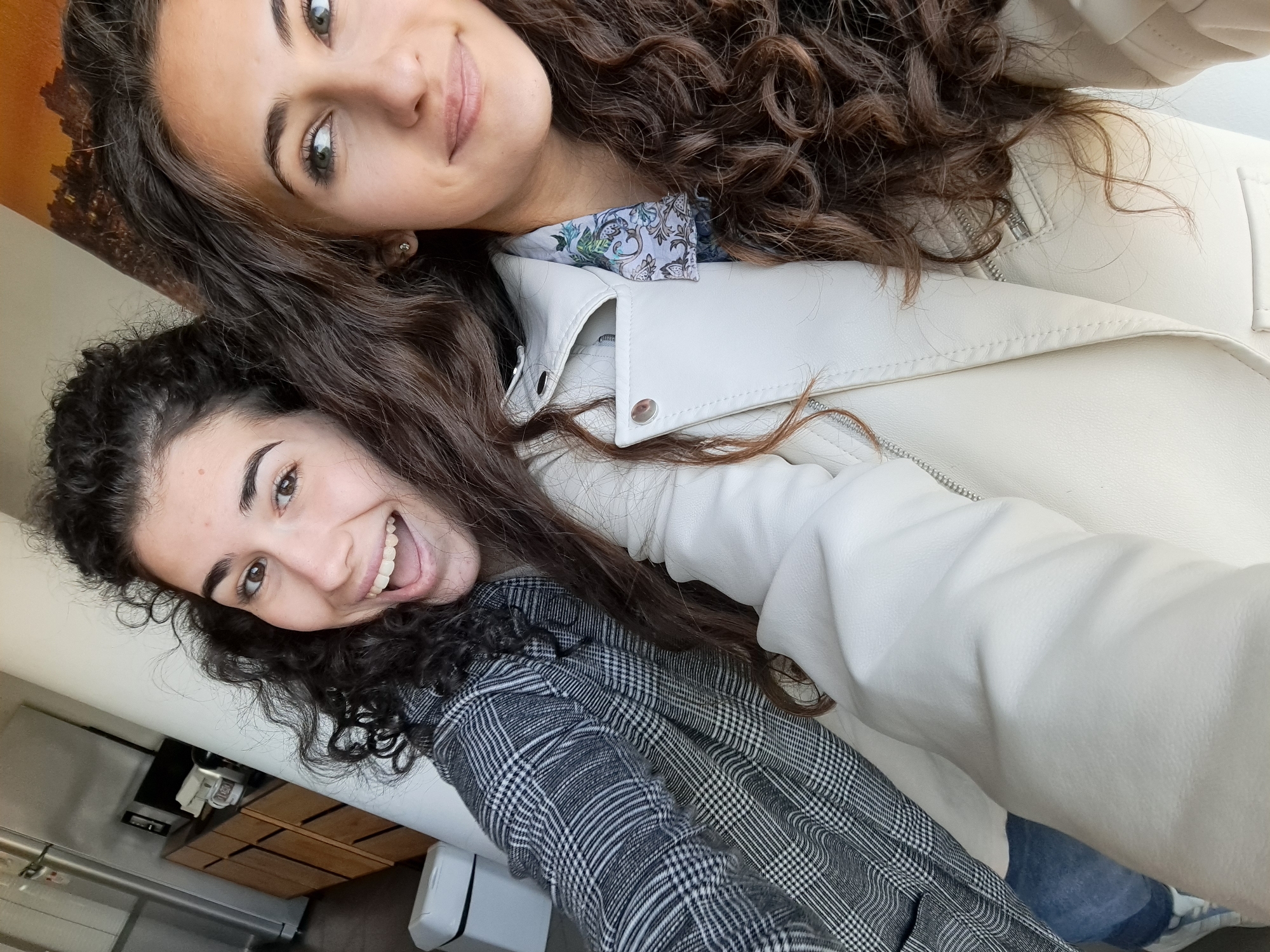 Two young women pose for a selfie. The woman at the top has long, dark, curly hair and is wearing a white jacket over a patterned shirt. The woman below also has dark, curly hair and is wearing a grey plaid jacket. The background shows a neutral-toned wall, and glimpses of what appears to be an interior. Based on visual analysis of the architectural style, it is not possible to confirm the location in Sofia, Bulgaria, or any other specific city without additional information.