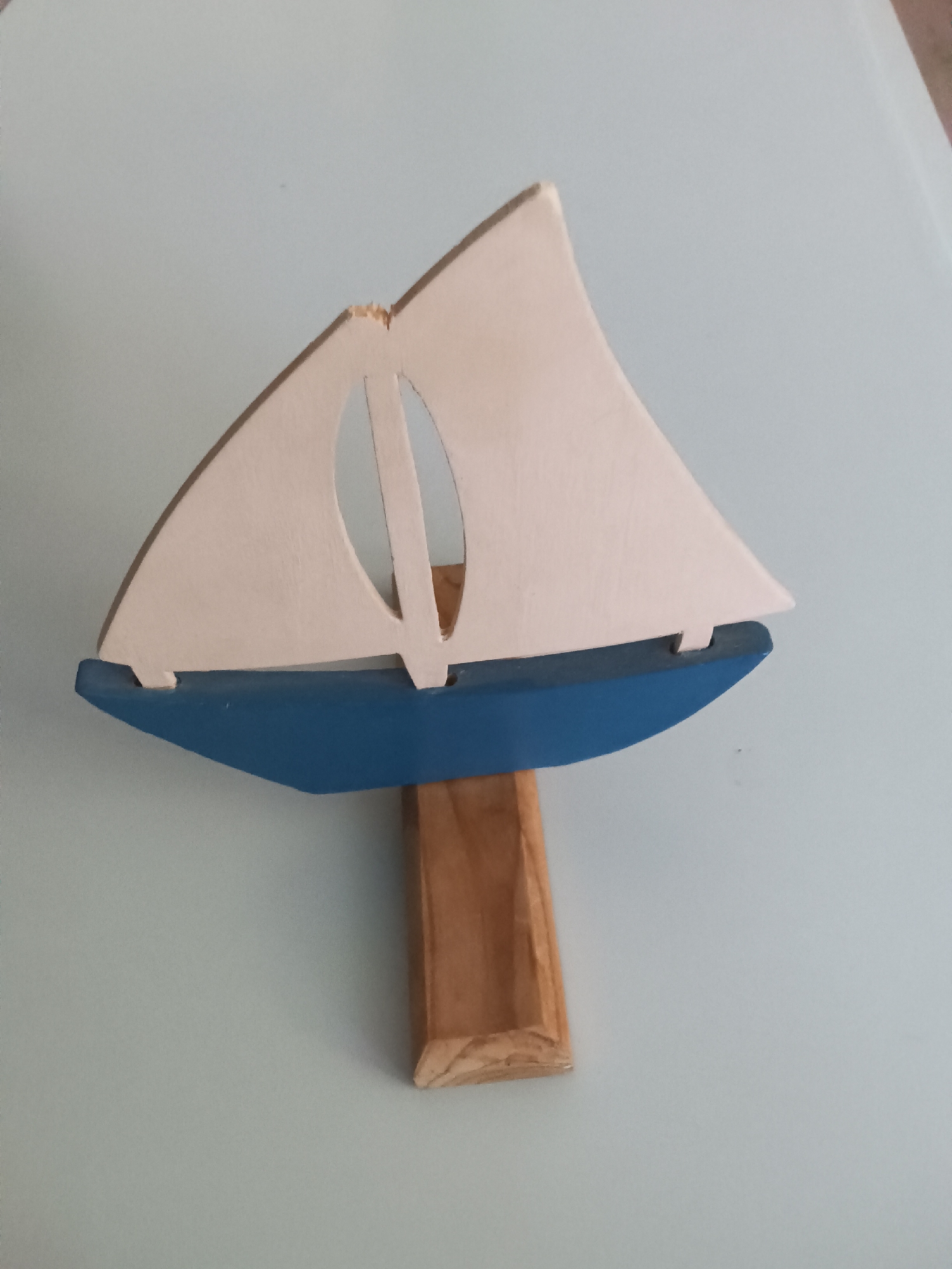 A wooden toy sailboat, painted blue with a white sail, is displayed on a light blue surface. The sailboat is mounted on a light brown wooden stand. The sail has an oval cutout and a small imperfection at the top. The background is a plain, light blue surface. The location in Makkum, Netherlands is not discernible from the image.