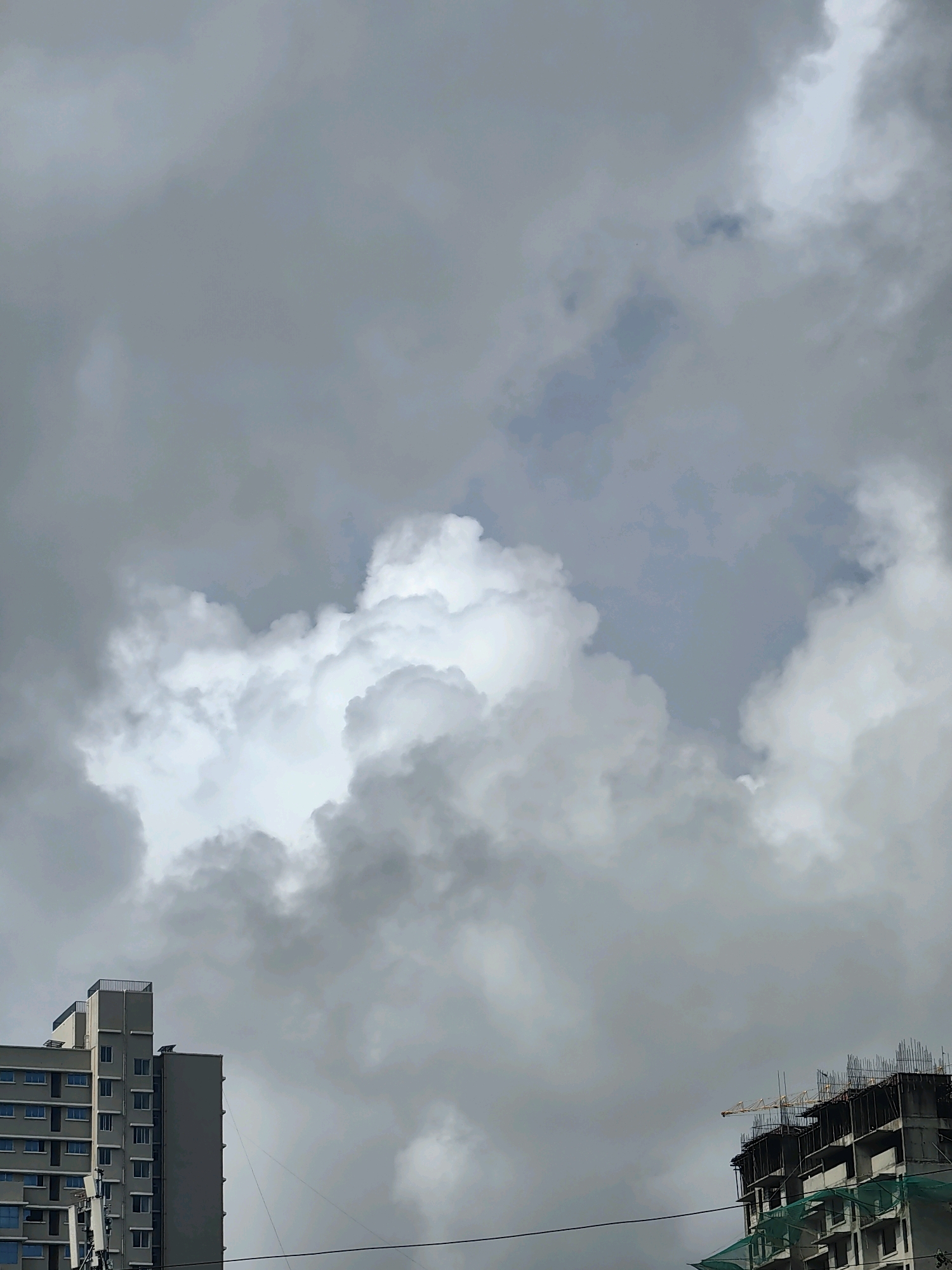 A low-angle shot captures a cloudy sky above two buildings in Mumbai, India. On the left, a completed, gray, multi-story residential building with numerous windows is visible. On the right, a concrete building under construction, with exposed rebar and green netting, is topped by a yellow crane. The sky is filled with a mix of dark and light gray clouds, with a patch of blue visible in the upper center. The image appears to be taken during the daytime.