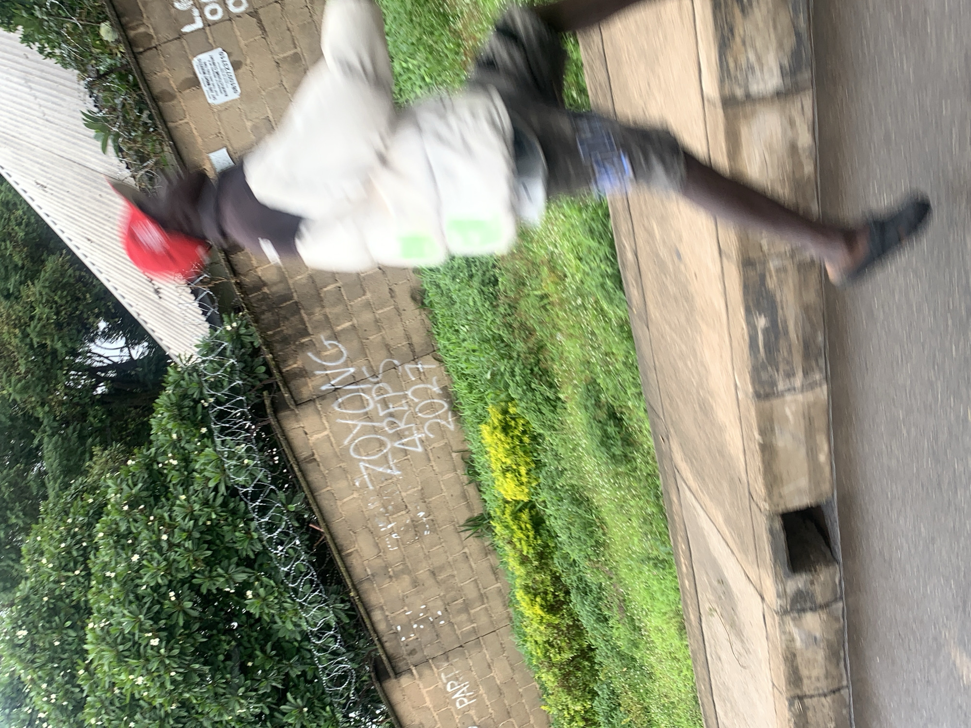 The image, captured in Jos, Nigeria, shows a blurred figure of a person running alongside a sidewalk. The person is wearing a red cap, a white puffer jacket over a dark hooded garment, and patterned shorts. They are also wearing dark slip-on shoes. In the background, a brick wall is visible, adorned with graffiti that includes the words "ZOYONG", "4PEPS", and "2027", as well as "PART". Barbed wire runs along the top of the wall, and lush green vegetation, including flowering bushes, grows behind it. The sidewalk has a drainage grate. The image is taken from a low angle, implying the photographer may be on the road.
