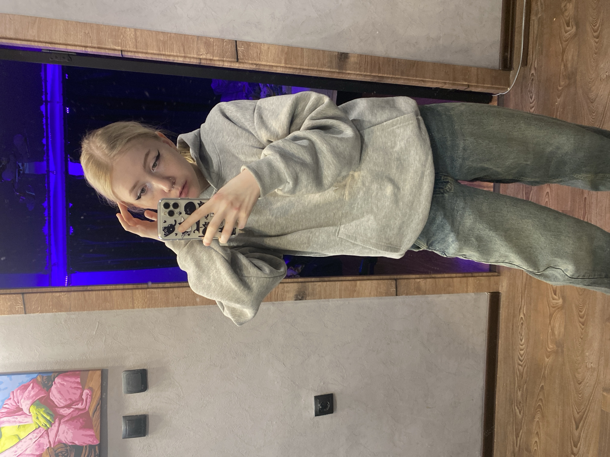 A young woman with blonde hair is taking a selfie in a mirror. She is wearing a gray hoodie and jeans. The mirror reflects a room with wooden floors and gray walls. There is a painting on the wall, along with some electrical outlets and switches. The location may be in Izhevsk, Russia, but the specific details to confirm this are not visible in the image.