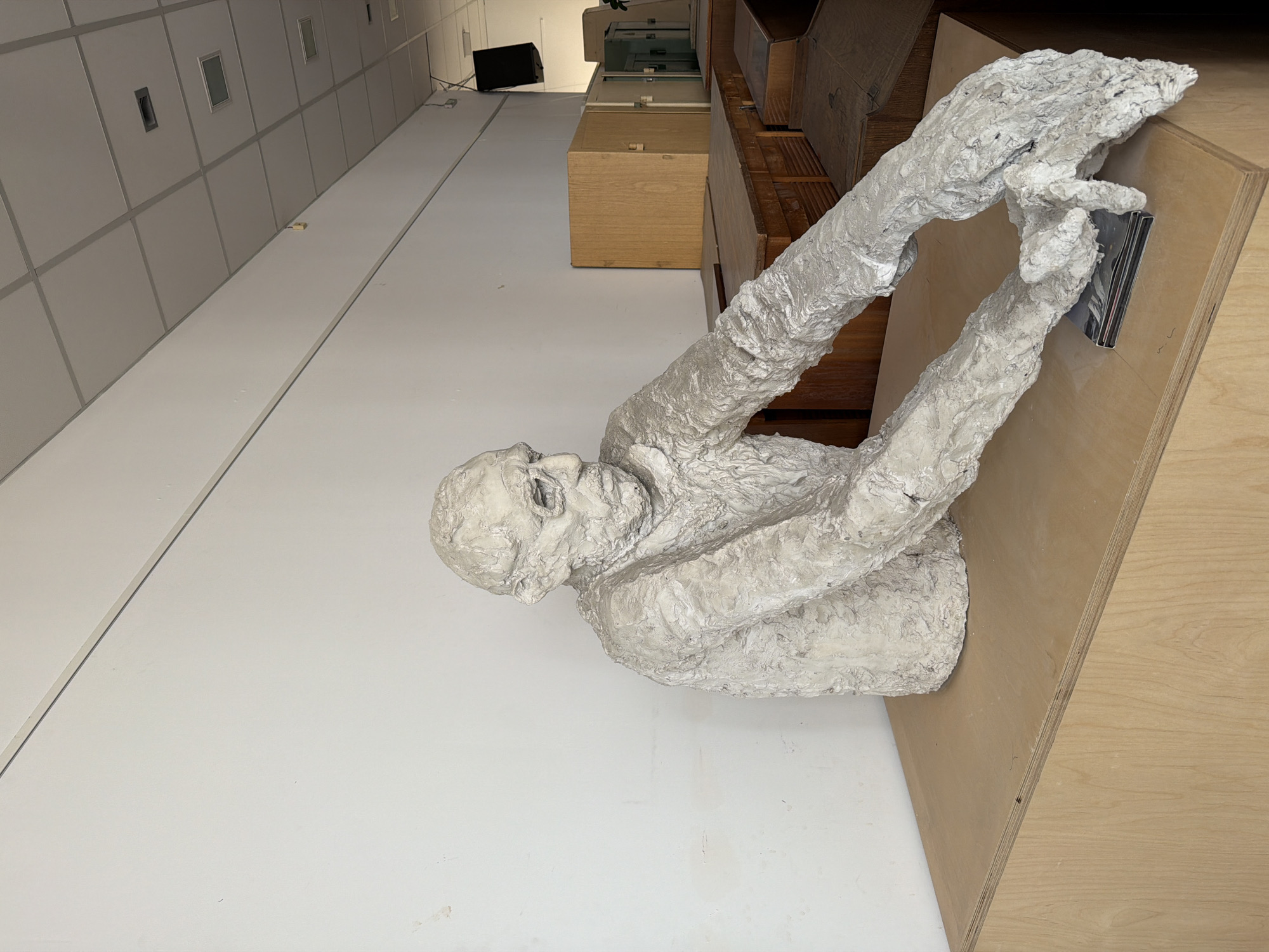 A low-angle, eye-level shot captures a white-painted plaster sculpture of a seated human figure, resting its chin on its chest with arms raised in front of it. The sculpture is holding a small, metallic object in its hands. The sculpture is positioned on a wooden surface with visible wood grain. The background includes a white tiled wall with small rectangular cutouts, and a series of wooden cabinets and boxes. Further in the background, a black electronic device is visible, possibly a monitor. The location is possibly in a public or gallery space in Plovdiv, Bulgaria.