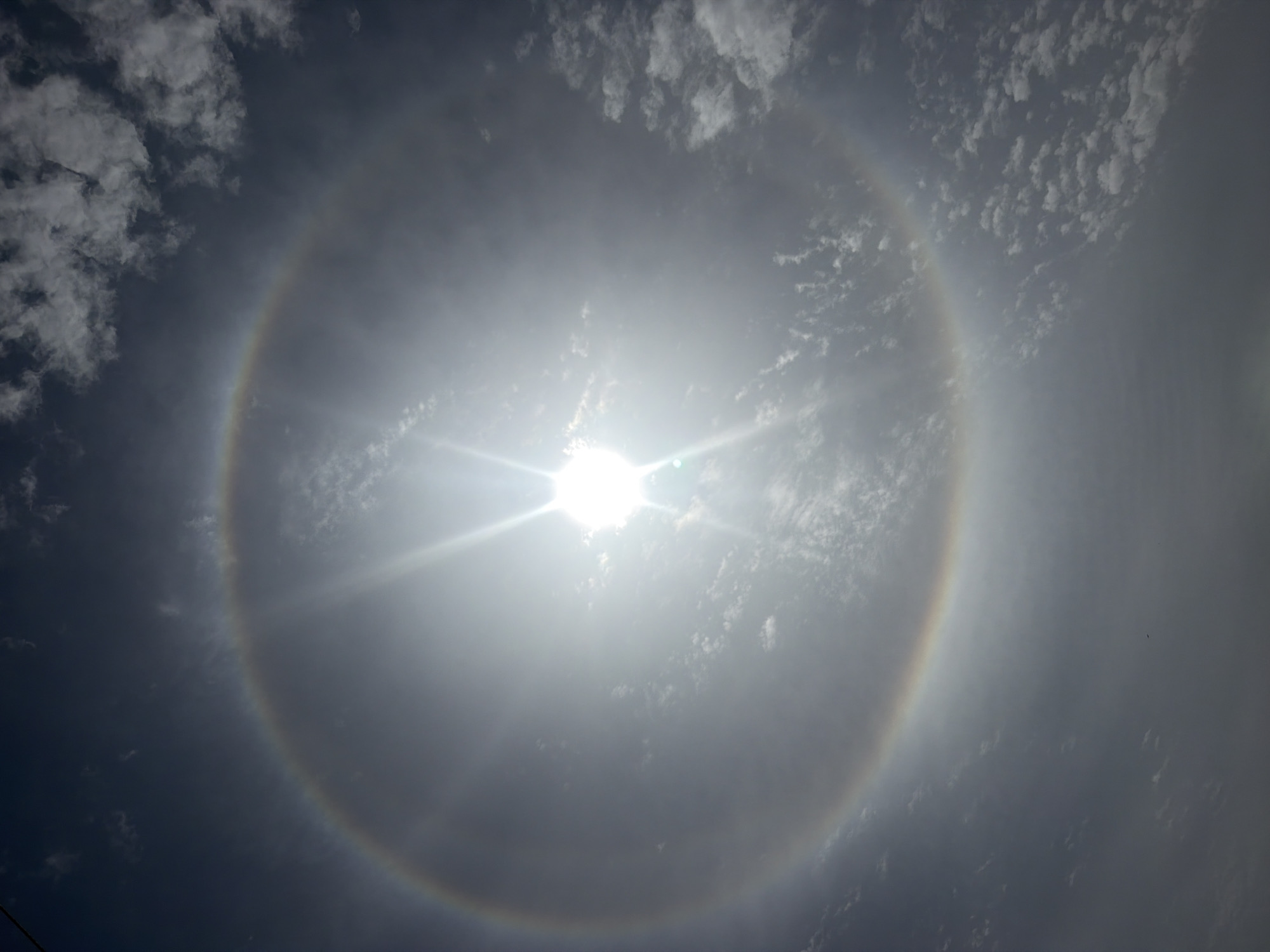 This image captures a sun halo phenomenon over Bukuru, Nigeria. The sun, a bright white disc with prominent rays, is centrally located in the frame. Encircling the sun is a distinct, luminous ring, exhibiting faint prismatic colors, most notably visible as a rainbow arc at the bottom of the halo. The sky is a gradient of dark grey at the top to a lighter, hazy grey towards the bottom. Scattered, wispy white clouds are present throughout the sky, some appearing dense and others more translucent. The overall lighting is bright due to the sun's intensity. There are no people or man-made structures clearly identifiable in this atmospheric scene.
