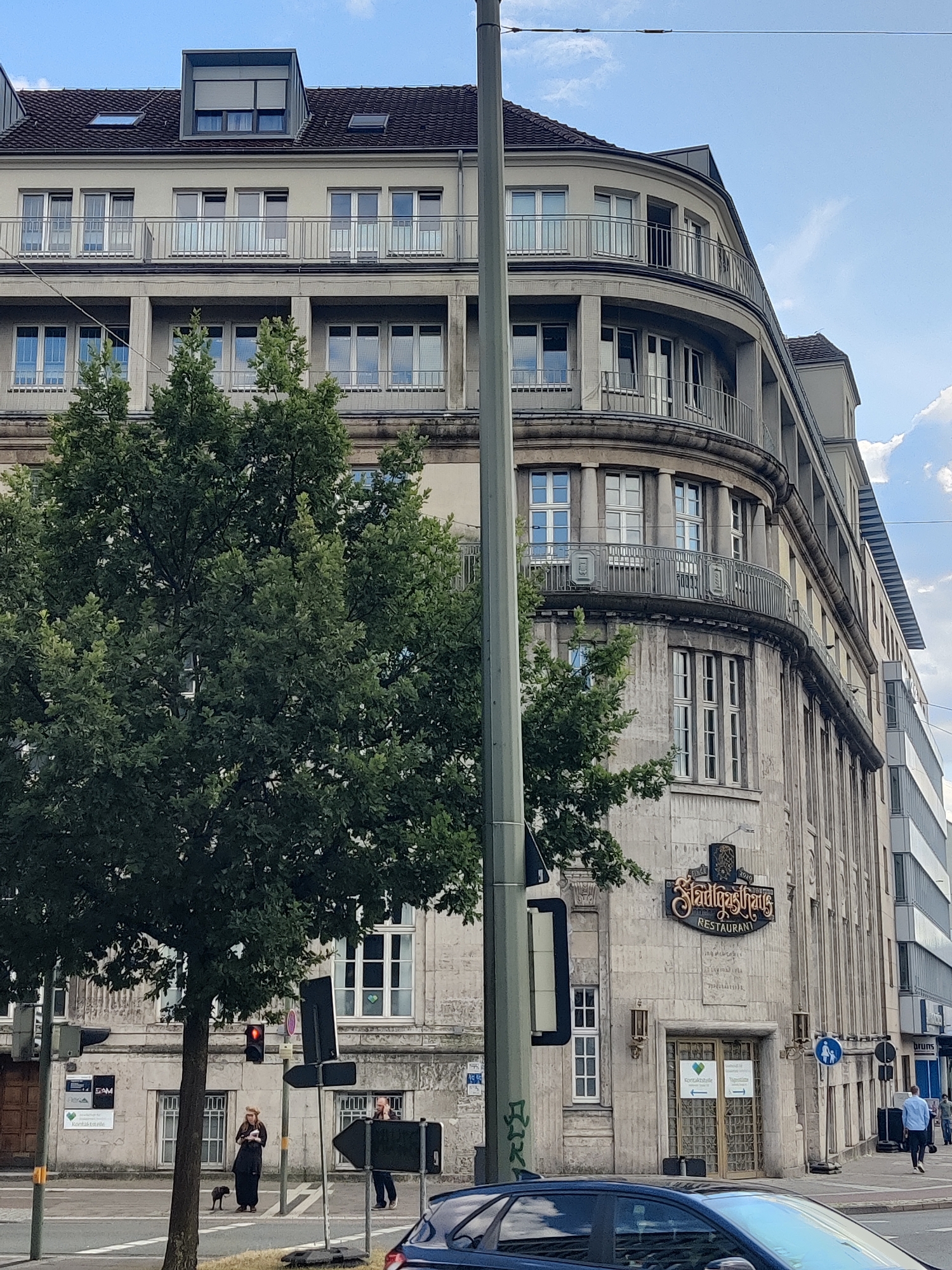 The image shows the facade of the "Stadthaus" building in Bielefeld, Germany, on a sunny day with scattered clouds. The building features a distinctive rounded corner and balconies with ornate railings. A large tree with dense green foliage is situated in the foreground, partially obscuring the lower floors of the building. On the ground level, a restaurant with the name "Stadthaus Restaurant" is visible above the entrance. To the left of the entrance, there are posters on the wall, and a traffic light shows red. A woman in dark clothing and a hat stands on the sidewalk with a small dog. A man is also visible further down the street on the right, walking away from the camera. A dark-colored car is parked in the lower right corner of the frame. The overall scene depicts an urban environment with architectural elements characteristic of early 20th-century design.