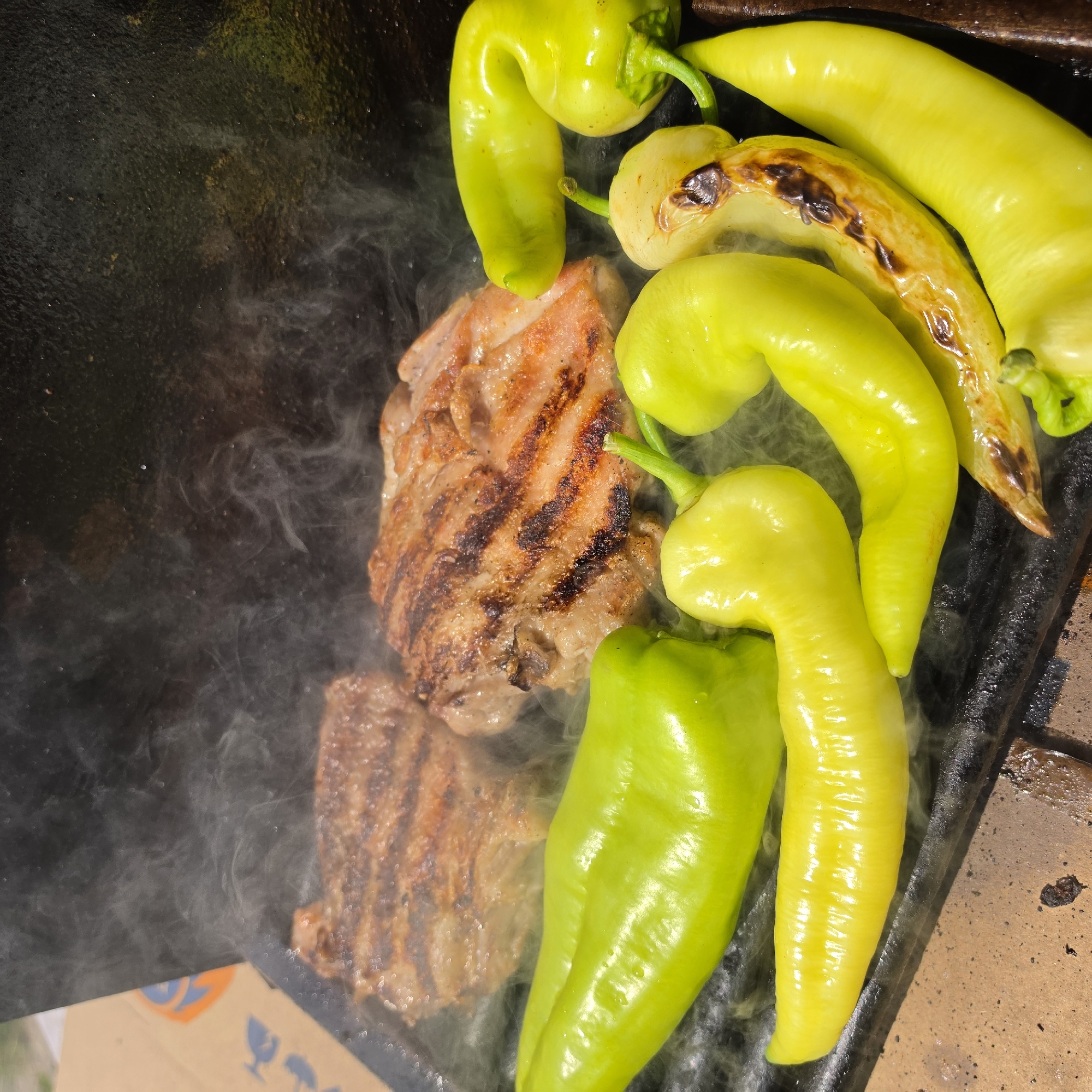 The image shows a close-up of two pieces of meat being grilled on a flat-top grill, accompanied by several bright green and yellow peppers. Wisps of steam rise from the hot grill, indicating the food is actively cooking. The meat appears to have grill marks and is browned. The peppers are mostly whole, with some showing slight charring from the heat. The grill surface is dark and slightly textured, set against a background that suggests an outdoor or rustic setting, possibly a brick surface. This scene is located in Peshtera, Bulgaria.
