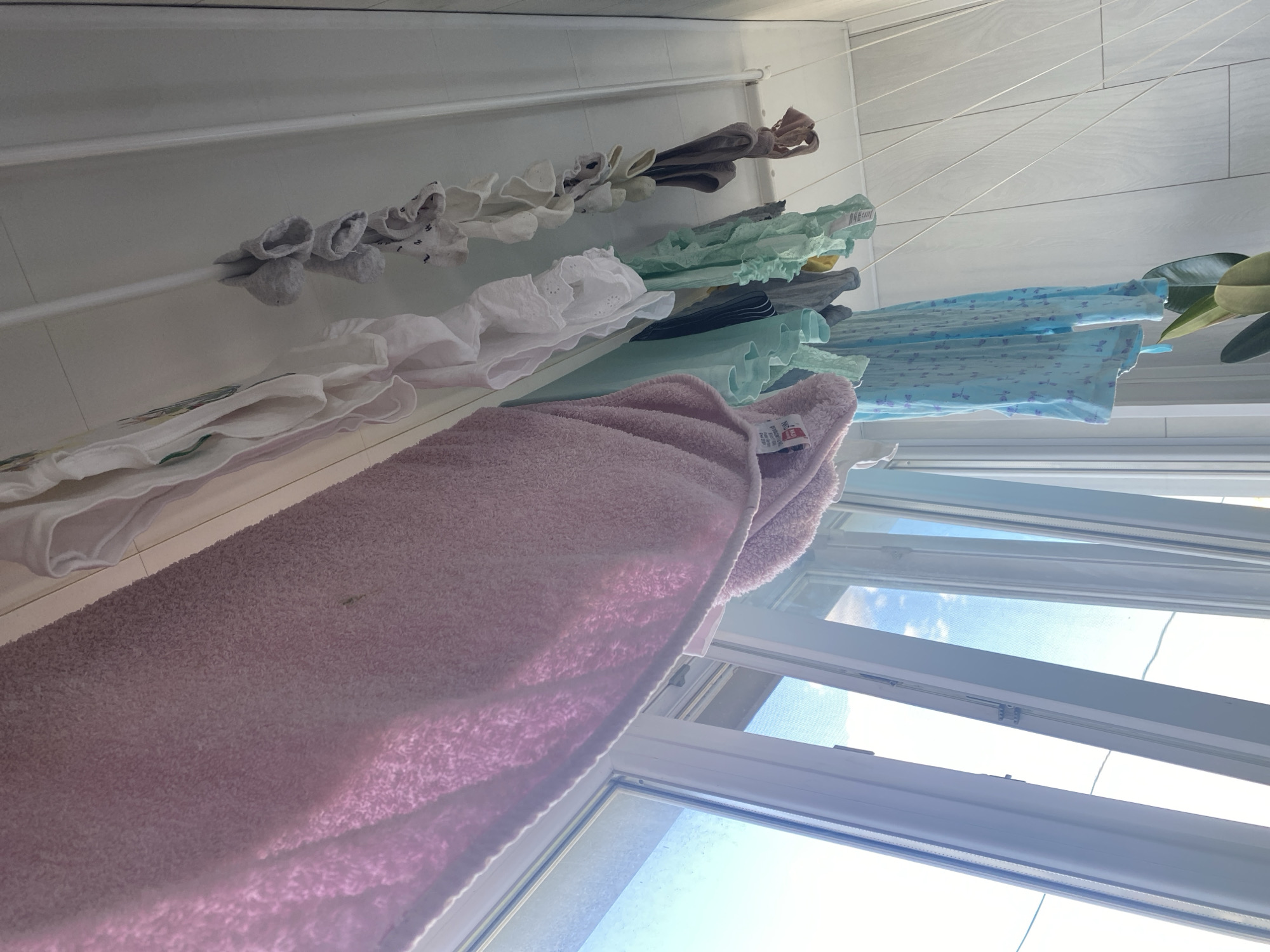 A clothesline is set up near a window in what appears to be a residential building. Various items of laundry are hanging on the line, including a large pink towel, several pairs of socks, and other garments in pastel colors like blue and mint green. The window is open, allowing sunlight to stream in, illuminating the laundry and the interior space. Outside the window, a bright sky with some clouds is visible. The walls inside are white and paneled. A plant with large green leaves is positioned at the bottom right of the frame. The scene conveys a sense of domesticity and everyday life, likely in a home in Odesa, Ukraine.