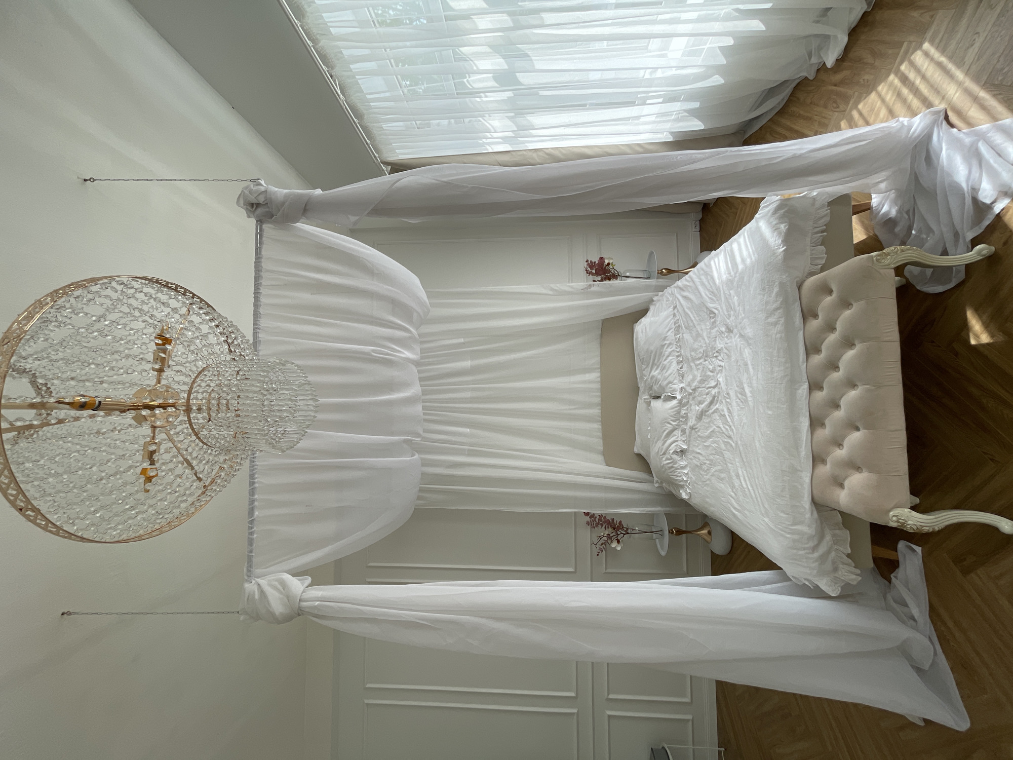 A slightly high-angle indoor shot shows a white canopy bed in what is likely a hotel room in Kazan, Russia, since it looks like a hotel bedroom. The bed is upholstered with white fabric on the headboard and on the bedding. Two small white tables, each with a small vase with pink flowers, stand on each side of the headboard. A crystal chandelier hangs at the left of the canopy. The canopy is draped in white fabric. A white curtain is in the background near a window. The floor is parquet. The walls are white and decorated with panels.