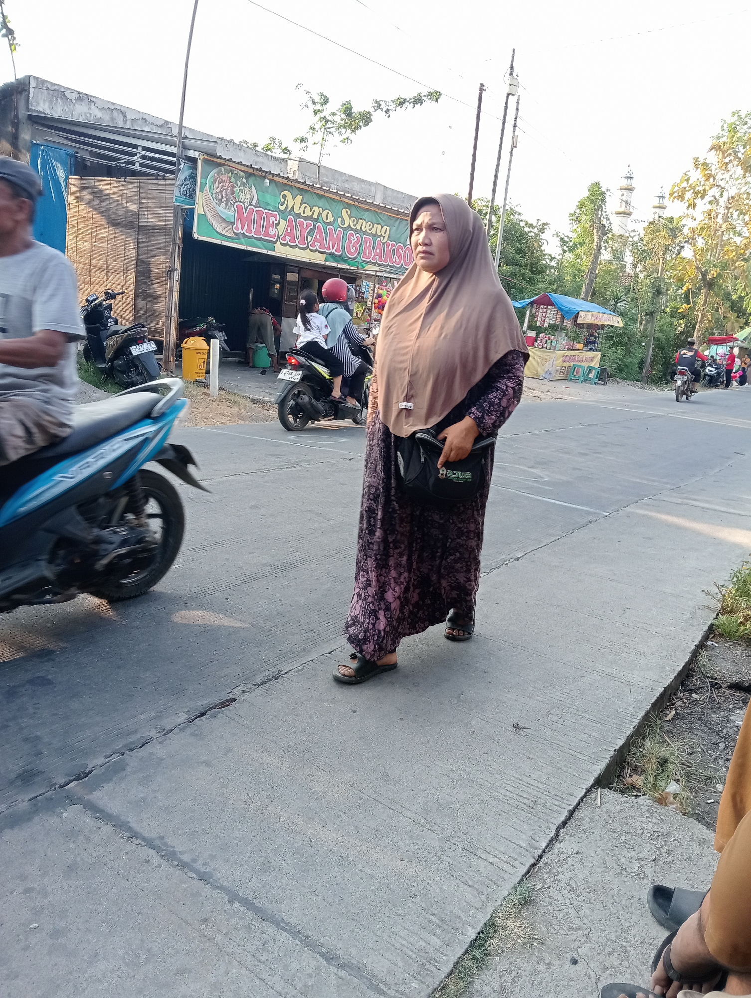 A middle-aged Indonesian woman, wearing a brown hijab and a floral patterned dress, walks on a sidewalk. She is carrying a black shoulder bag. To her left, a blue and black motorcycle is passing by. Behind her, a building with a sign that reads "Moro Seneng Mie Ayam & Bakso" is visible, suggesting it is a food establishment. In the background, there are more buildings, trees, and a mosque with minarets, indicating a likely urban or semi-urban setting in Tanjunganyar, Indonesia. The sky is bright and clear.