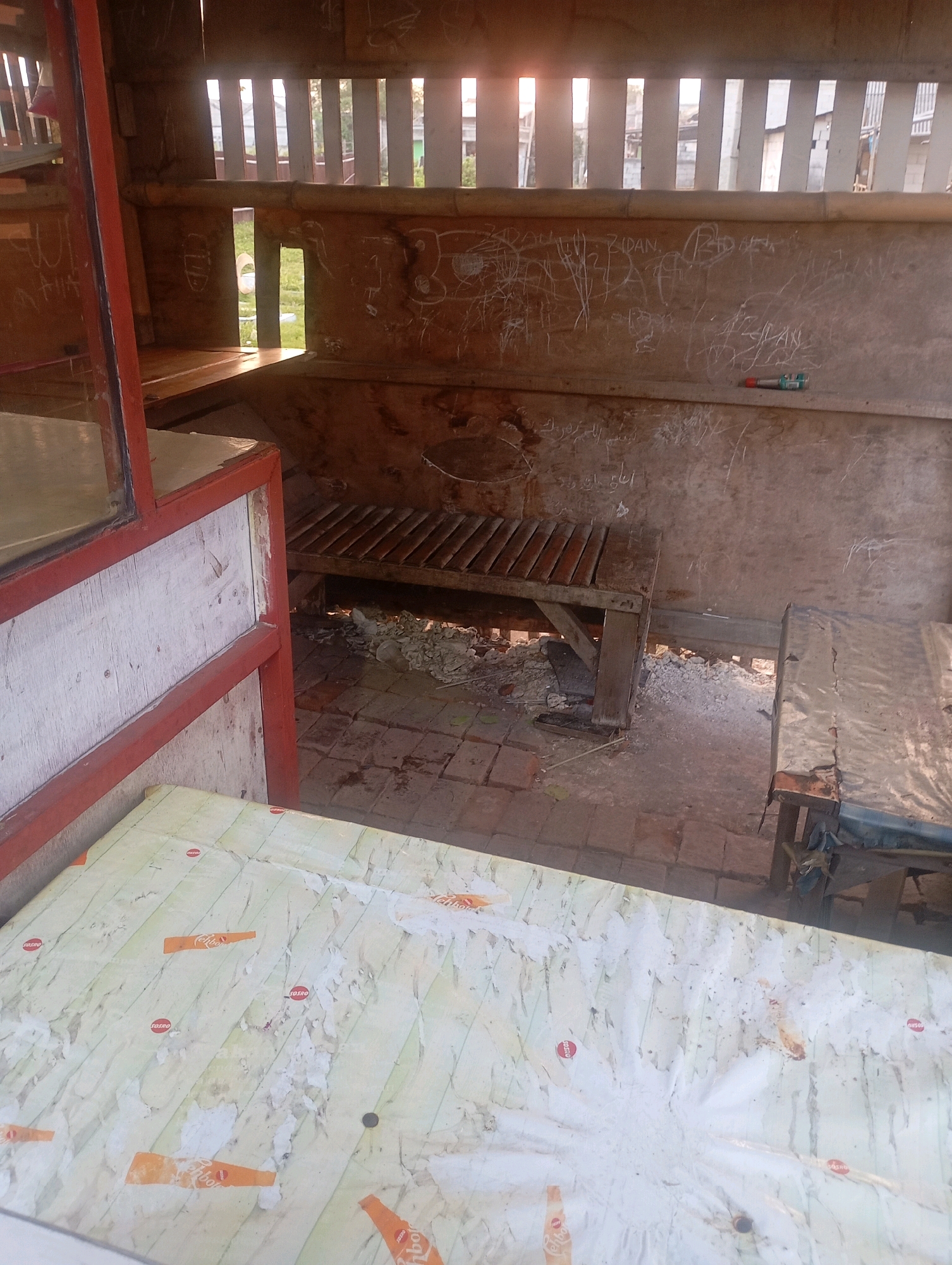 The image shows the interior of a small, open-sided structure, likely a roadside stall or shelter, in Babelan, Indonesia. The walls are constructed of rough wood and covered in various graffiti. Two wooden benches are visible within the structure, one appearing to be a simple slatted bench and another covered with a patterned sheet. The ground is paved with bricks and shows signs of disrepair, with debris scattered around. In the foreground, a table or surface is covered with a light-colored, patterned sheet, possibly a tablecloth or protective covering, with some torn paper remnants. Sunlight streams through vertical slats in the rear wall, illuminating the interior. A pen is visible on the wall in the middle ground. The overall scene suggests a rustic and possibly abandoned or underutilized space.