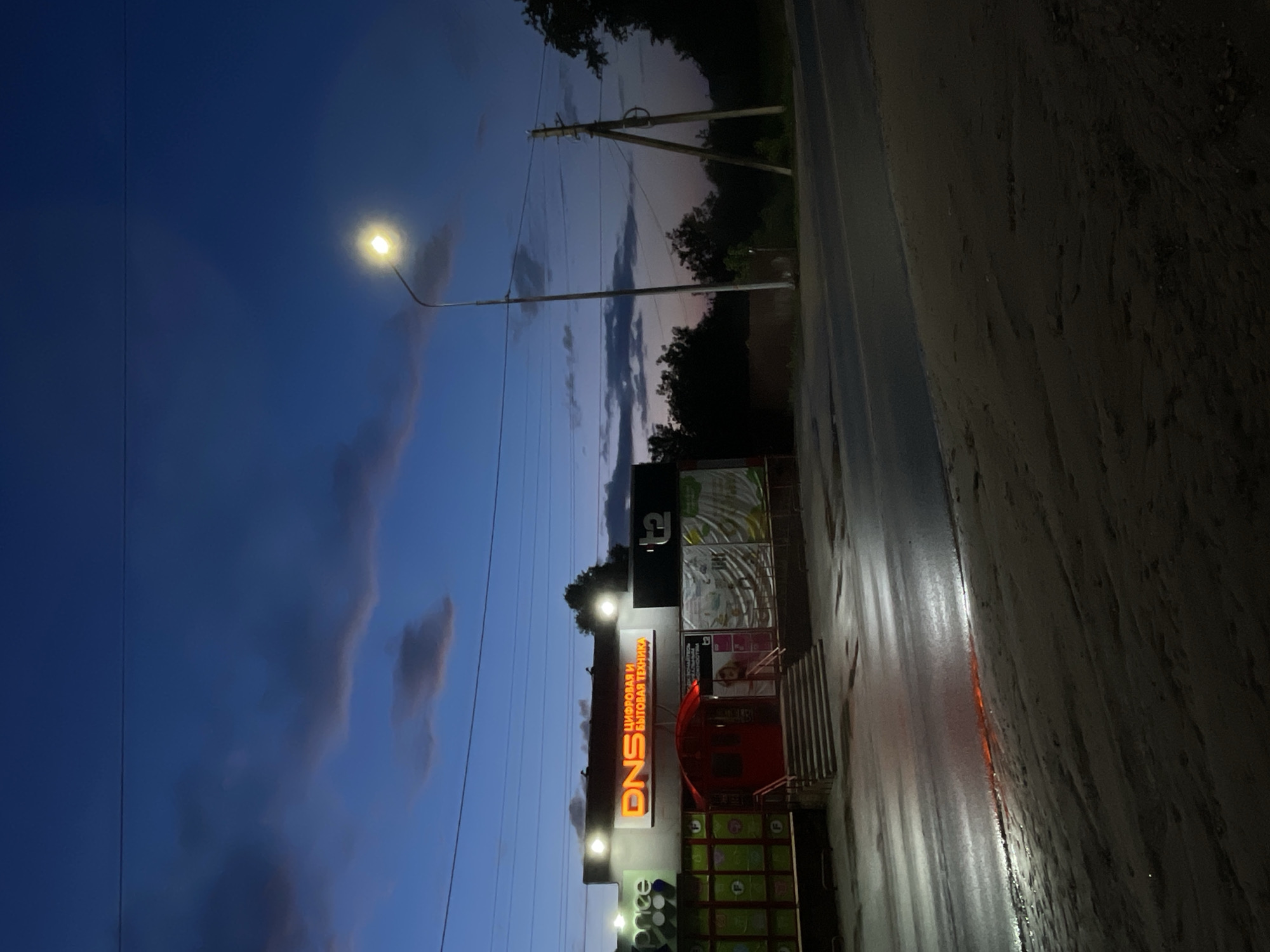 The image presents a nighttime or late dusk scene in Красные Баки, Russia. The sky, a deep indigo blue, features scattered clouds with remnants of an orange and pink sunset near the horizon. A bright, glowing streetlight on a tall pole illuminates the upper left portion of the frame. Power lines and utility poles are visible against the sky, extending towards the right.
In the foreground, a wet road or pavement surface reflects the artificial lights and the sky, suggesting recent rain or high humidity. On the right, a commercial building stands prominently, adorned with several illuminated signs. The most striking sign, in glowing orange letters, displays "DNS". Directly above it, smaller orange text reads "ЦИФРОВАЯ И БЫТОВАЯ ТЕХНИКА," which translates to "DIGITAL AND HOME APPLIANCES." Other less discernible signs, including a "t2" logo and faint "price" text, are also present on the building's facade. A red entrance door with steps leads into the building. The background reveals dark silhouettes of trees and foliage. The overall atmosphere is quiet and calm, characteristic of an evening street scene.