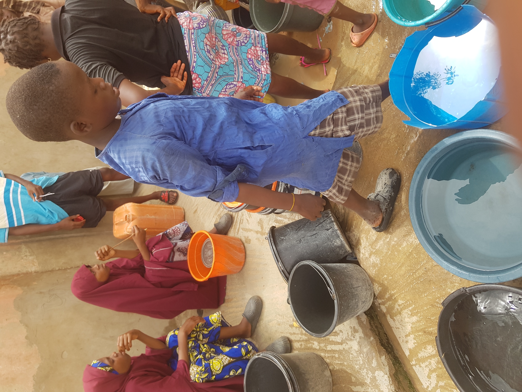 In Dutsinma A, Nigeria, a group of people stand and sit near an assortment of buckets, barrels, and containers, many of which are filled with water. A young boy in a blue shirt and plaid shorts is walking. Several of the individuals are women, some wearing maroon hijabs and colorful patterned dresses. The ground appears to be wet and possibly muddy.