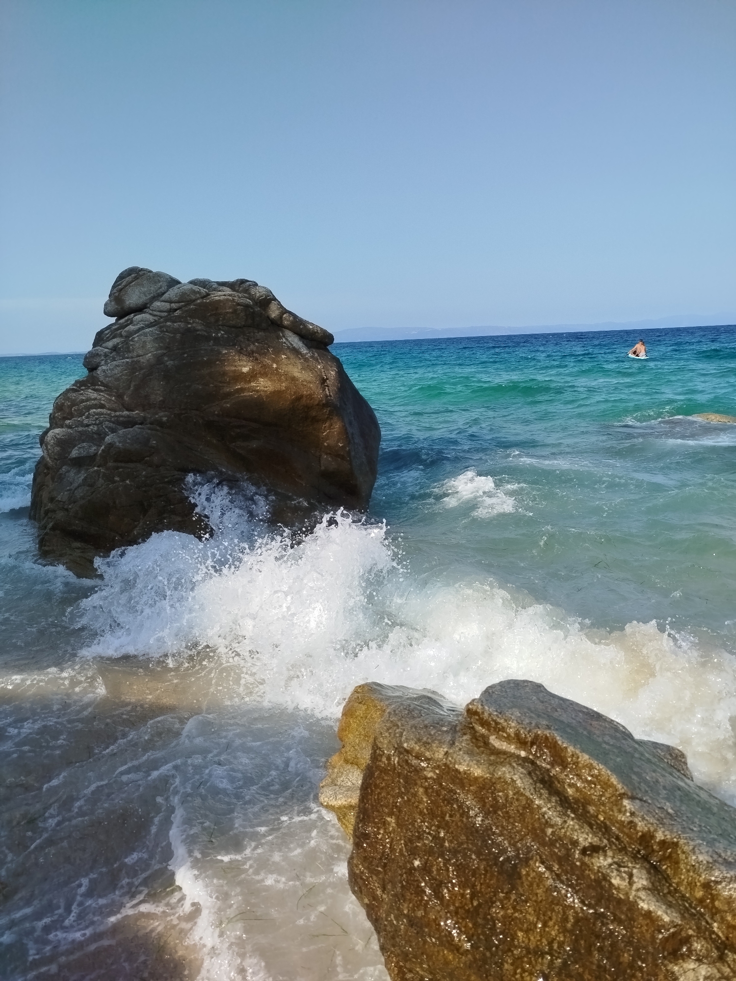 The image depicts a rocky beach scene in Sithonia, Greece. A large, dark boulder is positioned in the foreground, partially submerged in the turquoise water. Waves are crashing against the boulder, creating white foam and spray. In the background, the clear blue sky meets the horizon, where a faint line of land is visible. Further out in the water, a person is seen floating on their back, appearing relaxed. The overall impression is one of a serene, natural coastal landscape.