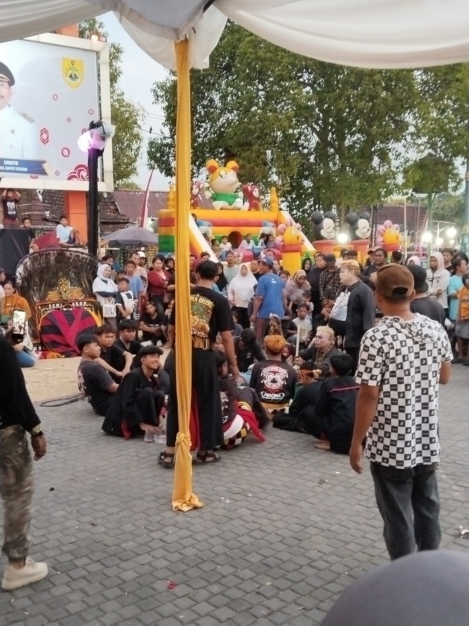 The image captures an outdoor event in Sragen, Indonesia, with a crowd gathered around a performance area. In the background, a large inflatable structure resembling a bouncy castle with Mickey Mouse characters and other cartoon figures is visible. Above the crowd, a large banner displays a portrait of a man in official attire, with text in Indonesian, likely a local official.
The foreground features several individuals in traditional attire, appearing to be part of a performance or ceremony. Some are seated on the ground, while others stand. A prominent yellow fabric hangs from above, adding a decorative element to the scene. The ground is paved with interlocking bricks. The overall atmosphere suggests a festive or cultural gathering.