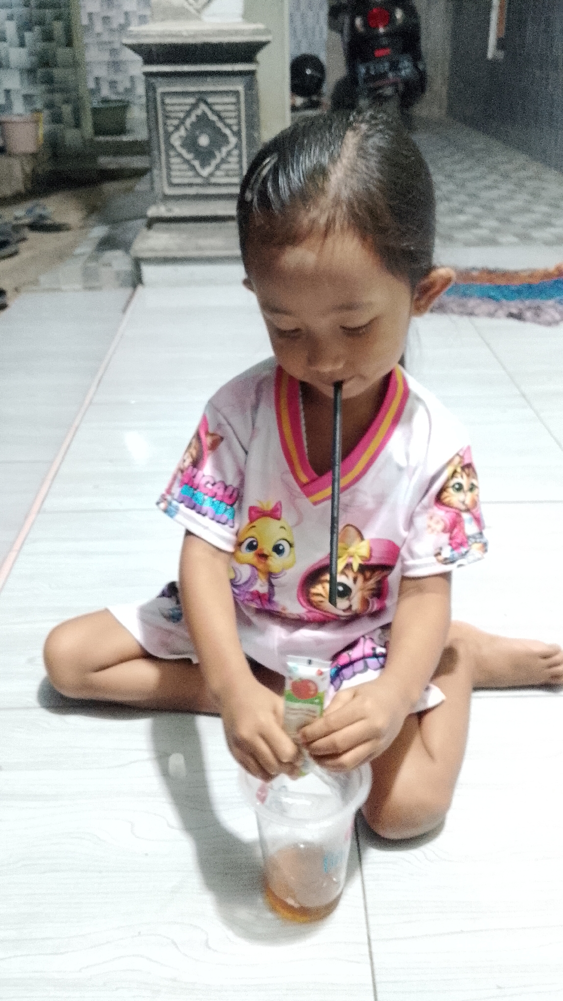 In Seputih, Indonesia, a young girl sits cross-legged on a tiled floor, focused on a plastic cup containing a brown liquid. She wears a white t-shirt with a v-neck and colorful cartoon prints of animated characters. A black straw extends from the cup to her mouth. Her hands are engaged with a small, foil-pouched packet, seemingly adding its contents to the drink. The background suggests an outdoor or semi-outdoor setting, with a textured pillar, a motorcycle, and a mat visible. The lighting is dim, creating shadows on the floor.