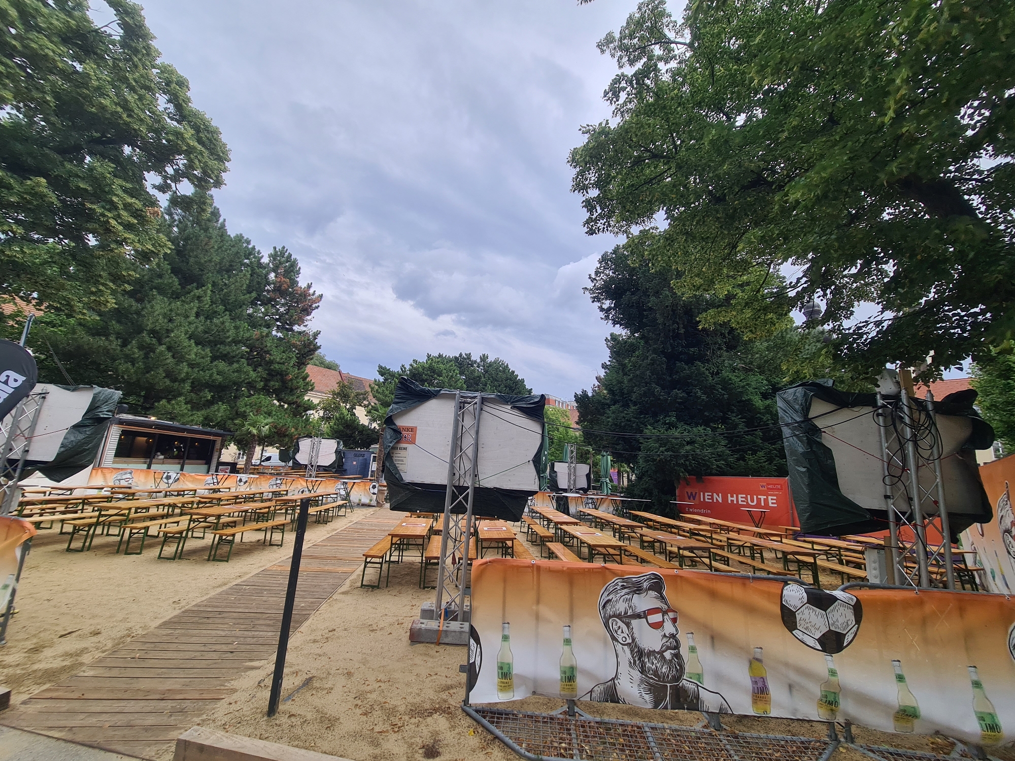 This image shows an outdoor seating area with picnic tables and benches set up on sand. The area appears to be prepared for an event, possibly a fan zone for a sporting event, given the presence of a soccer ball graphic on a banner. The banner also features an illustration of a man with a beard and sunglasses, along with bottles of a beverage. In the background, there are trees, buildings, and a cloudy sky. Signs in German are visible, including one that reads "WIEN HEUTE," suggesting the location is in Vienna, Austria. The scene is empty of people.
