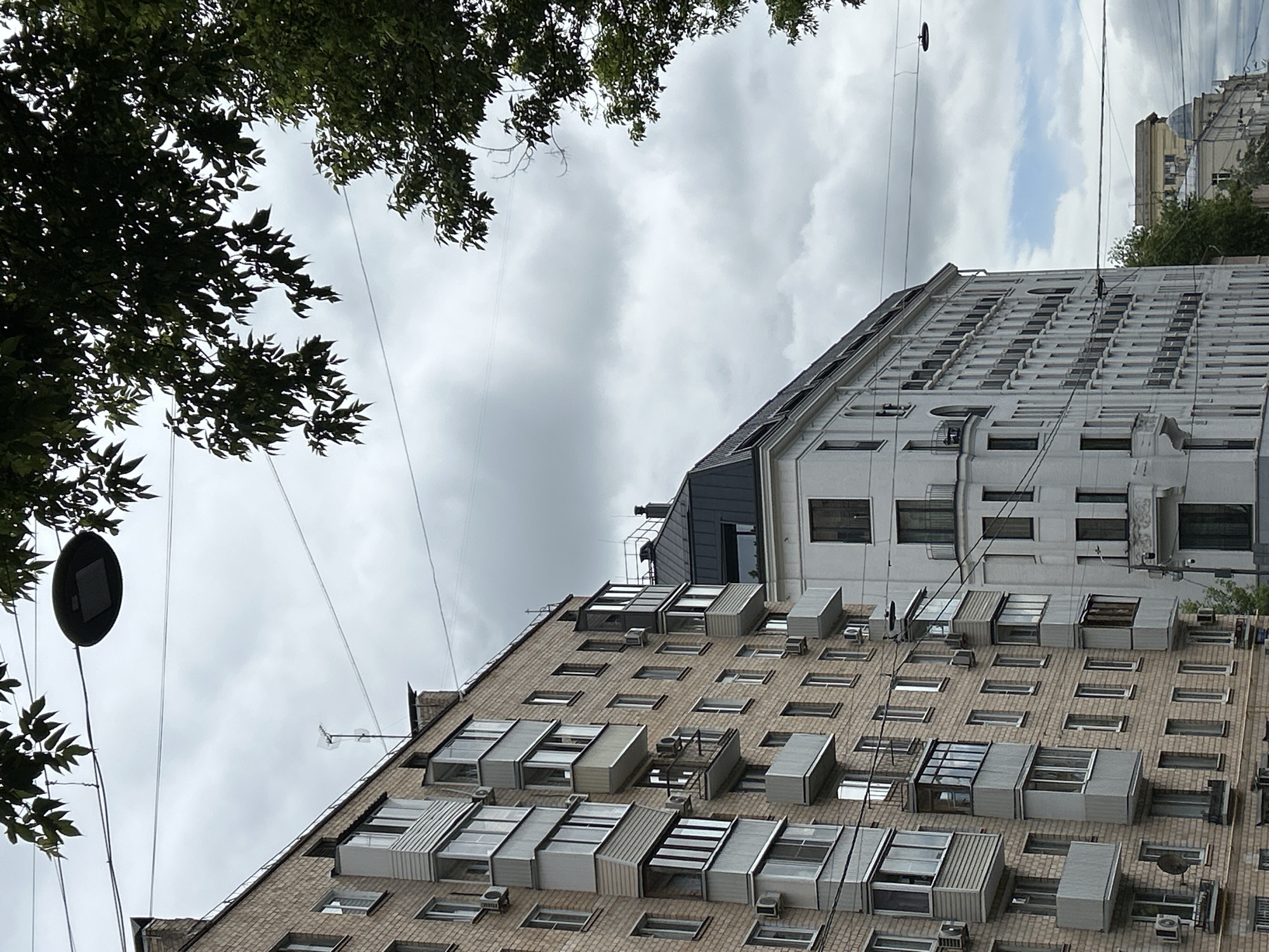 The image shows a view looking up at two buildings in an urban environment. The building in the foreground is constructed of brick and has many windows, most of which have protruding metal frames. Several air conditioning units are visible below the windows. The building in the background is white with a grid-like pattern of windows. Power lines extend across the sky. The sky is overcast with grey clouds. Trees with green leaves are visible in the upper left corner and partially obscure the view. There is a round, black object attached to a pole on the left side of the frame. The city location is unknown.