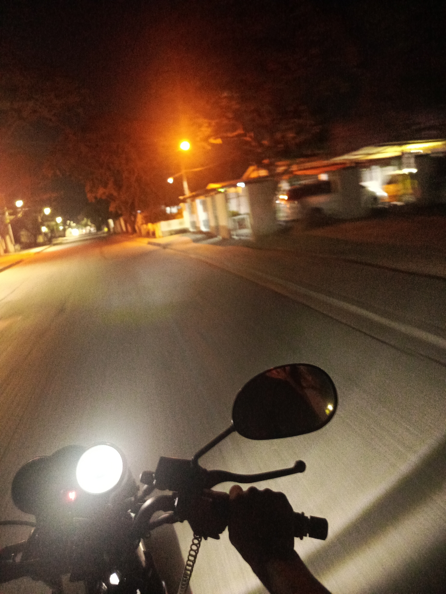 This nighttime image provides a first-person perspective from a motorcycle rider in Juan Griego, Venezuela. The bright, round headlight of the motorcycle dominates the lower foreground, illuminating the road directly in front of the vehicle and casting light on the handlebars. The rider's right hand, clad in a dark glove, is visible gripping the handlebar. A rearview mirror on the left side of the motorcycle reflects a partial, indistinct image, likely of the rider's head or face.
The scene depicts a street or road stretching into the distance, lined by trees and simple, low-rise buildings, possibly houses or small businesses, primarily on the right side. The sky is dark, with scattered, warm orange-yellow streetlights illuminating sections of the road and the surrounding structures. These lights cast long shadows and create an interplay of bright and dark areas. The road itself appears paved, with faint lines or markings visible further down the street. The background elements are slightly blurred, suggesting motion or a wide aperture in low light. The image captures the experience of navigating a street at night, relying on artificial light sources. No visible text is present in the image.