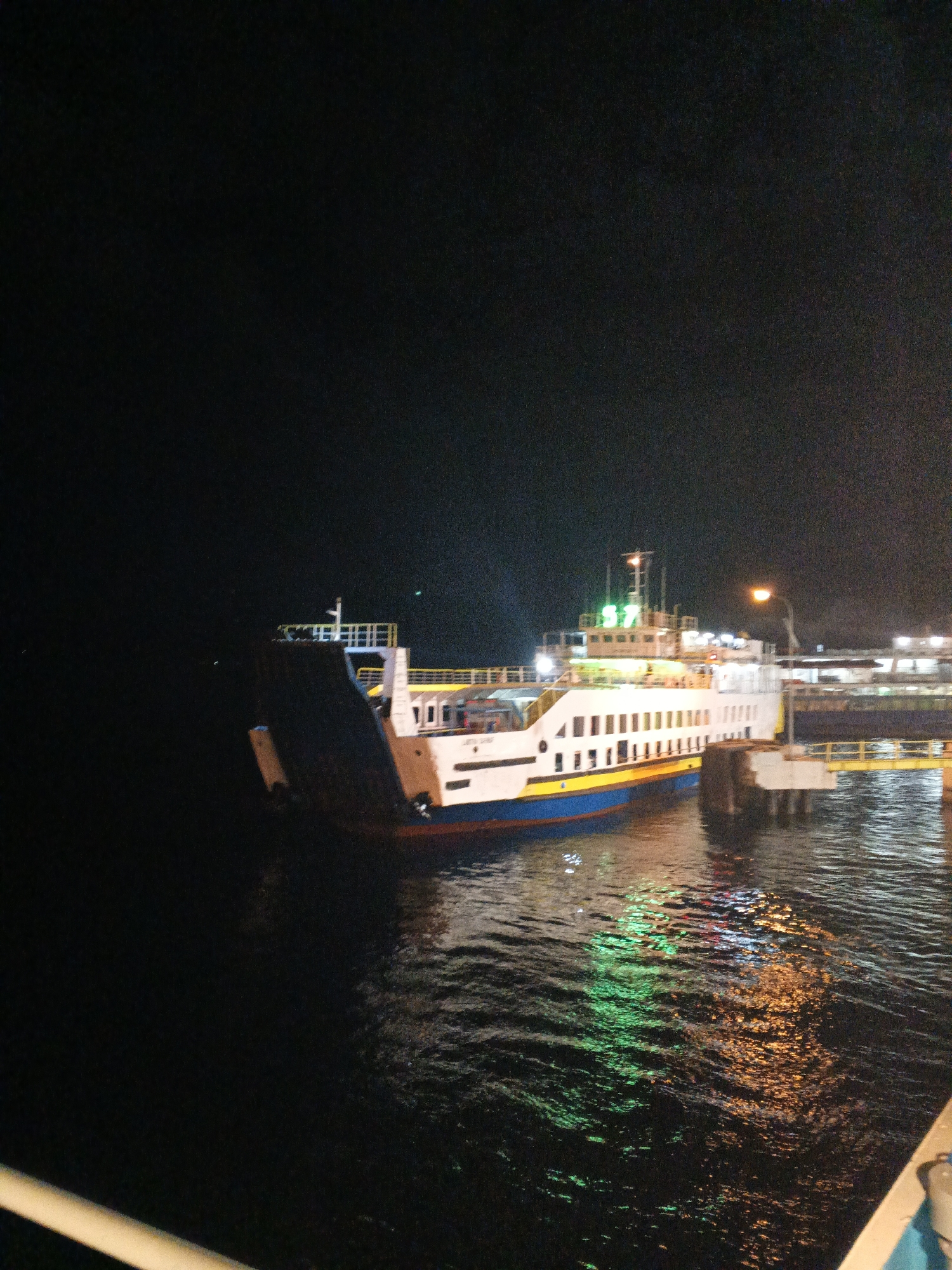 The image shows a ferry docked at a port at night, with the lights of the ferry and the port illuminating the scene. The ferry is white with a blue and yellow stripe along its hull. It has multiple decks and windows, and the number "57" is visible in green lights on its upper structure. The ferry is docked next to a concrete pier with yellow railings. The water in the harbor reflects the colorful lights of the ferry and the port. The background is dark, suggesting a clear night sky. The location is the port of Melaya, Indonesia. There are no people clearly visible in the image.