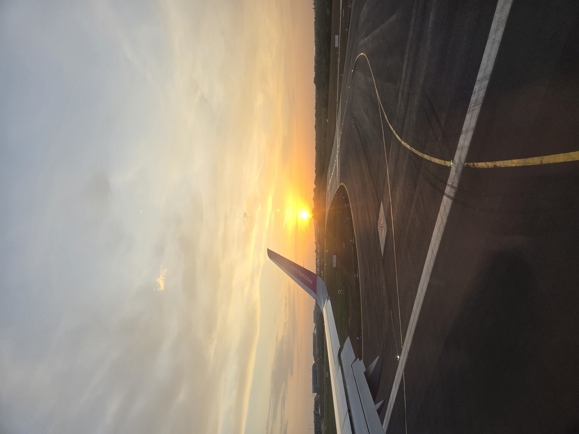 View from the interior of an aircraft during sunrise at an airport. The wing of the aircraft is visible. The aircraft is on the runway, indicated by painted lines. The sky is filled with clouds and the rising sun. The airport is near a forest. The aircraft belongs to the airline Wizz Air. The image does not contain sufficient information to accurately discern the location as North Hertfordshire.