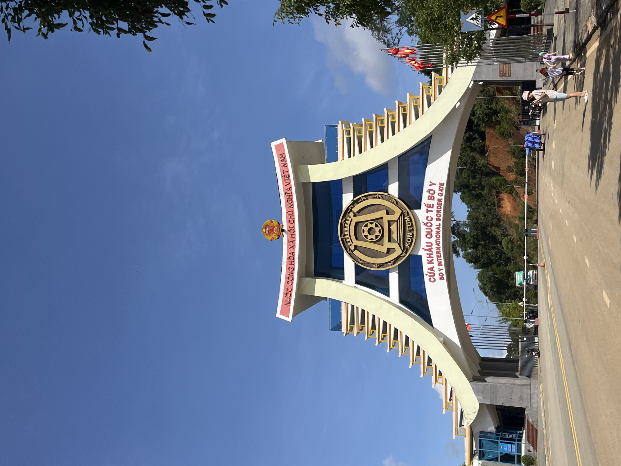 The image shows the Bo Y International Border Gate in Vietnam. The gate is a large, multi-story structure with a distinctive arched entrance adorned with Vietnamese national symbols and text in Vietnamese and English. Flags of Vietnam are prominently displayed on poles to the right of the entrance. The sky is clear and blue, with some trees and a hillside visible in the background.
In the foreground, there is a road leading to the border gate. Several people are visible, including a woman in a pink hat taking a photo, and two children walking away from the camera. There are also some vehicles, including a truck and motorcycles, suggesting active border crossing activity. The overall scene depicts a busy border crossing point under a bright, sunny sky.