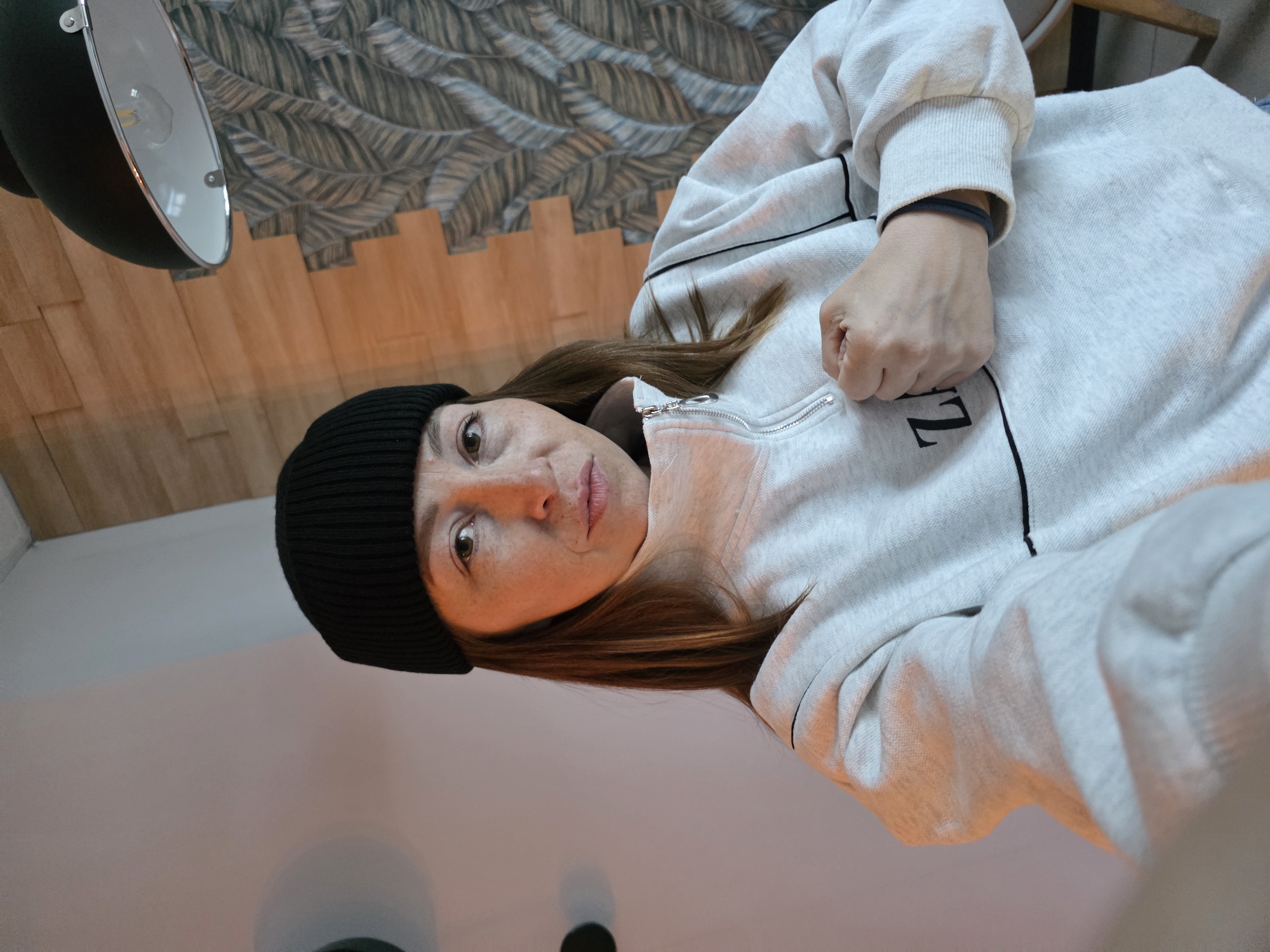 A medium shot depicts a woman wearing a black beanie and a gray sweatshirt with black trim. The woman is positioned so she is looking downward at the viewer, her head tilted slightly to the left. Her brown hair falls around her face, and she has freckles on her nose and cheeks. Her right hand is clenched into a fist.
Behind her, the wall is decorated with a pattern that resembles stylized leaves or feathers. A black and silver lamp is visible in the upper left corner of the image. The overall lighting suggests an indoor setting. The location is unspecified in the city of Unknown, Unknown.
