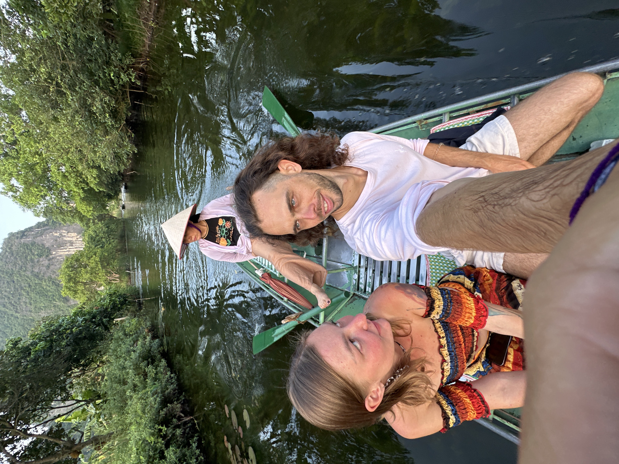 Three people are in a boat on a river. The person in the foreground on the left is a young woman with blonde hair, wearing a colorful off-the-shoulder top. The person in the foreground on the right is a young man with curly brown hair, wearing a pink long-sleeved shirt. The third person, in the background, is a woman wearing a conical hat and a pink jacket over a black top. The boat is green and appears to be made of metal. There are paddles in the boat. The river is dark and reflects the surrounding lush green trees and a large karst mountain in the background. The scene is likely located in the area of Tam Cốc-Bích Động, near Nam Hoa Lư Ward, Ninh Bình Province, Vietnam, a region known for its scenic river tours through karst landscapes. The overall lighting suggests it is daytime, possibly late afternoon or early morning, given the soft light.