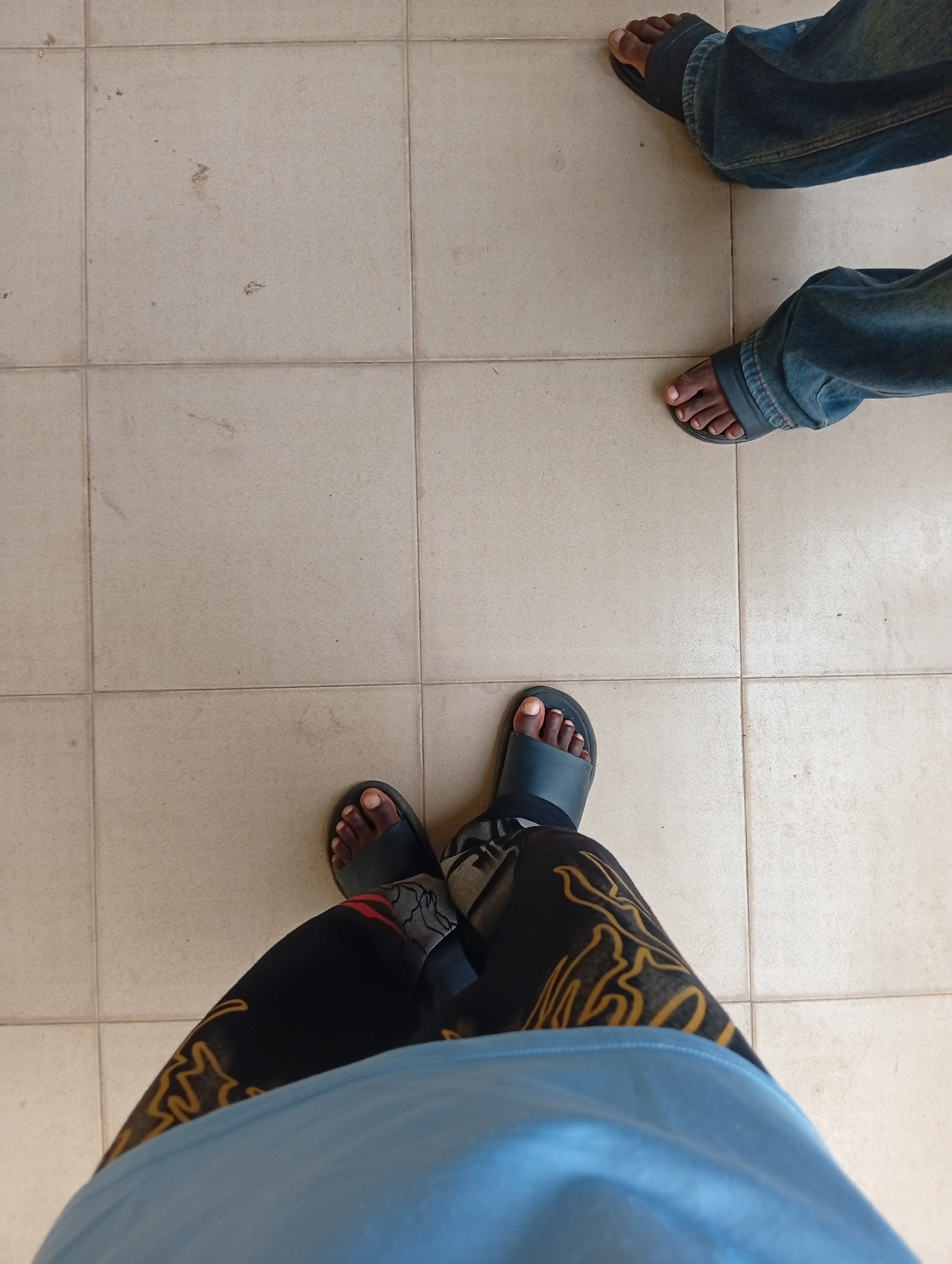 The image shows two pairs of legs and feet standing on a tiled floor. The floor is composed of light-colored square tiles, with visible grout lines.
The pair of legs on the left belongs to a person wearing a light blue garment on their upper body, black trousers with a yellow pattern, and dark blue open-toed sandals. Their feet are bare, and their toes are visible. The trousers have a patterned design that appears to be yellow outlines of figures or shapes.
The pair of legs on the right belongs to a person wearing blue denim jeans and dark blue open-toed sandals. Their feet are bare, and their toes are visible. The jeans appear to be slightly faded.
The scene suggests an indoor setting, possibly a hallway or public space, given the tiled floor. There are no other visible elements or interactions in the image. The lighting appears to be natural or ambient, casting soft shadows. The exact location in Ukukwa, Nigeria cannot be determined from the visual information in the image alone.