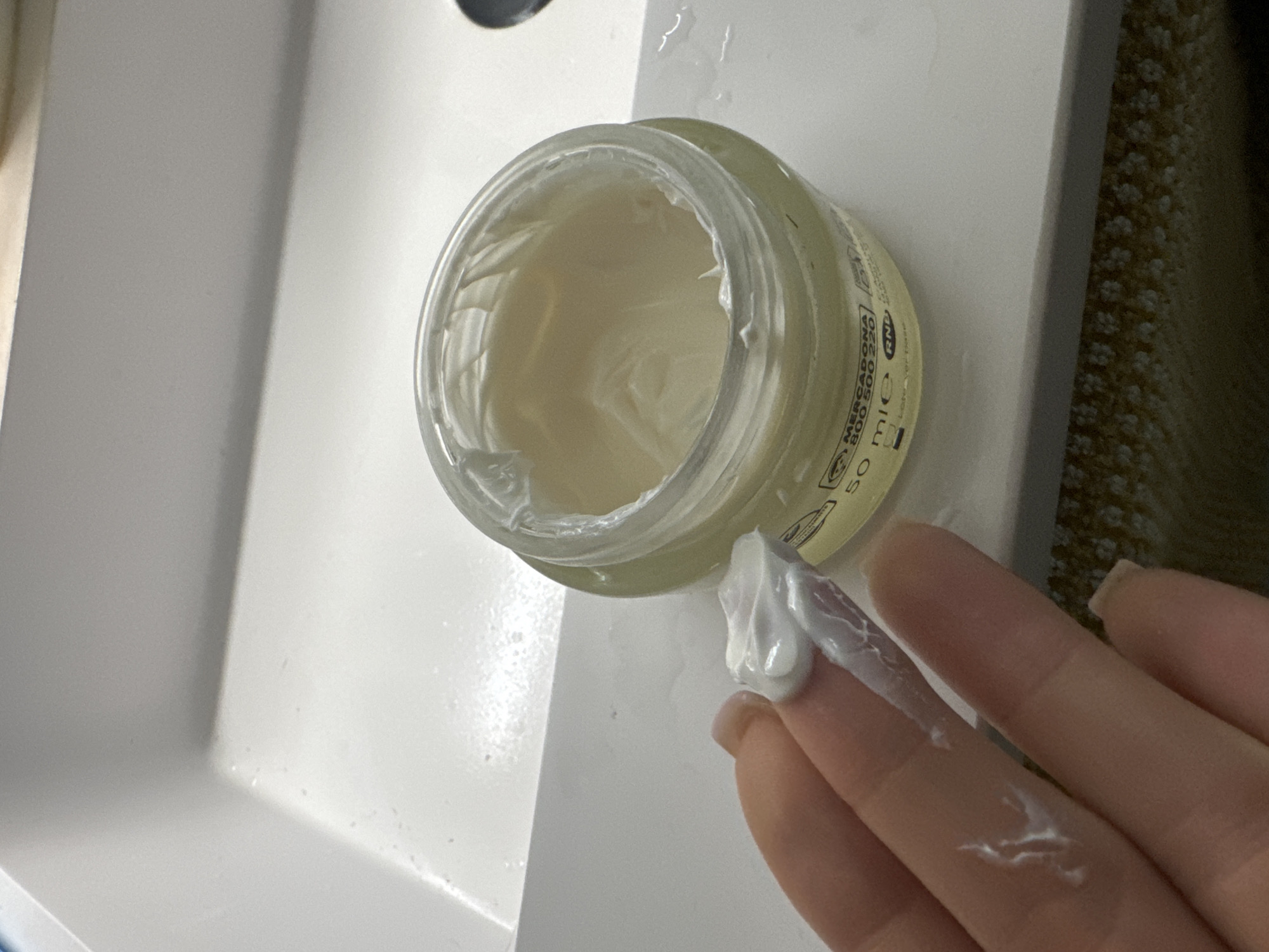 A hand with a dollop of white cream on its index finger rests above a clear glass jar of cream, which sits on a white countertop. The jar is open and the cream inside is also white. Text on the jar is partially visible, including "MERCADONA 800 500 220" and "50 ml e". A white sink is visible in the background, and a patterned rug is on the floor. The scene is indoors, likely in a bathroom. There is no information to indicate this scene is in Murcia, Spain.