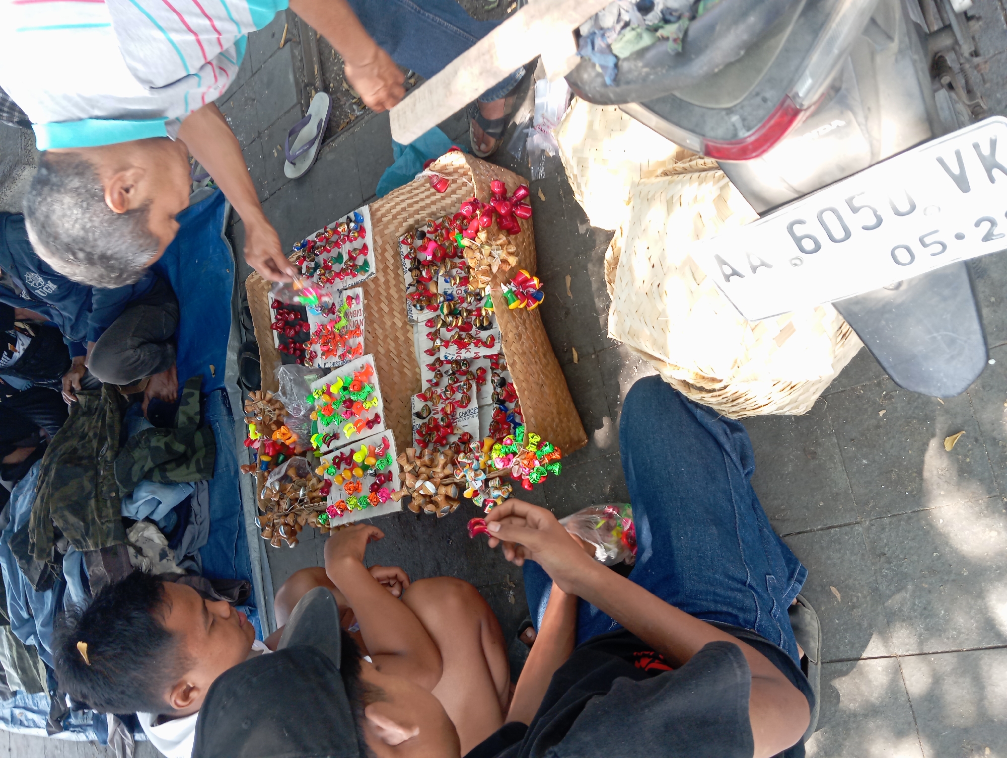 This image captures a street scene in Borobudur, Indonesia, likely near the famed temple complex. Two young men are seated on the ground, surrounded by an assortment of small, colorful trinkets displayed on woven mats and in a basket. One man, wearing a black t-shirt and jeans, holds a small plastic bag filled with similar items and appears to be examining them. The other young man, in a white hooded jacket and black baseball cap, looks down at the merchandise. An older man in a striped shirt leans over the display, seemingly interacting with the goods and possibly a customer. The ground is paved with square tiles, and a motorcycle is partially visible on the right, with its license plate reading "AA 6050 VK." The scene suggests a vibrant marketplace atmosphere, with vendors and potential shoppers engaging with handcrafted items. The background shows a blue tarp spread out, with various garments laid on top, indicating a setup for outdoor vending. The overall impression is one of a lively transaction occurring in a public, outdoor space within Borobudur.